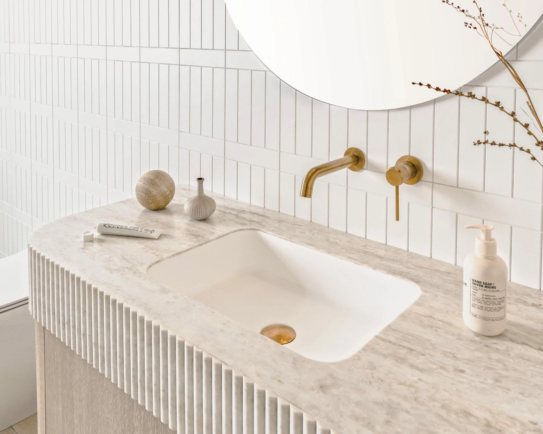 A modern bathroom sink area with a beige marble countertop, a white rectangular sink, and a gold faucet. Decor includes two small vases, a spherical object, a soap dispenser, and a mirror above the sink. The wall behind is covered in white tiles.