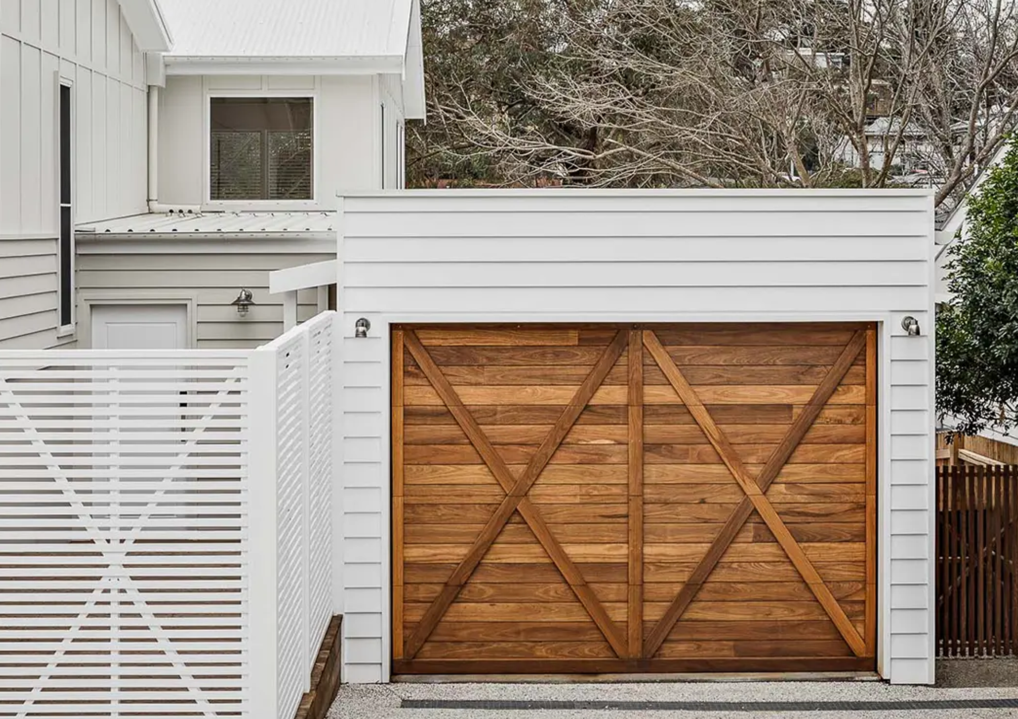 A wooden garage door with cross-bracing, attached to a white house with siding, next to a white fence and trees in the background.