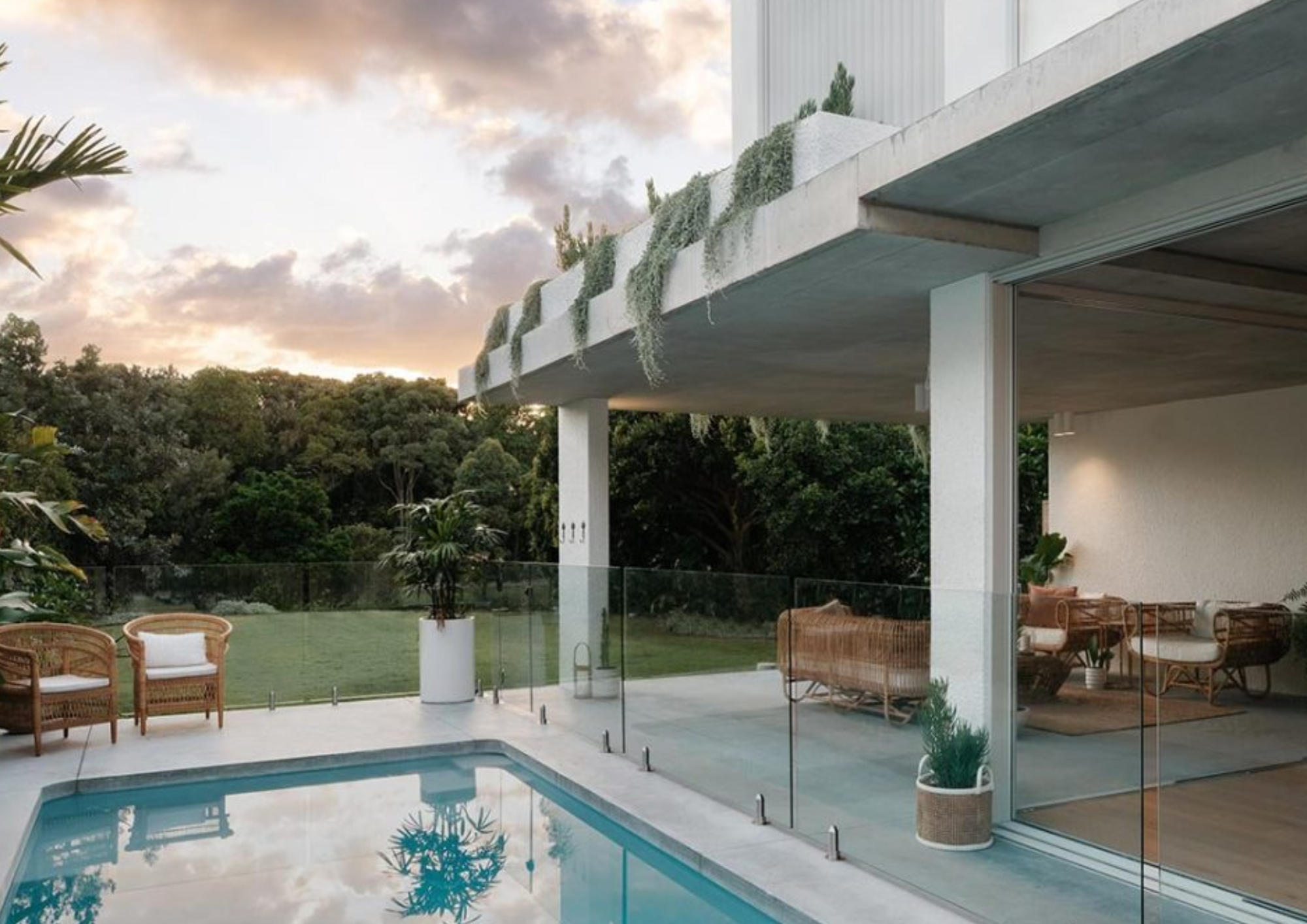 Modern outdoor patio with a swimming pool, wicker chairs with white cushions, potted plants, and glass fencing, overlooking a lush green landscape during sunset.