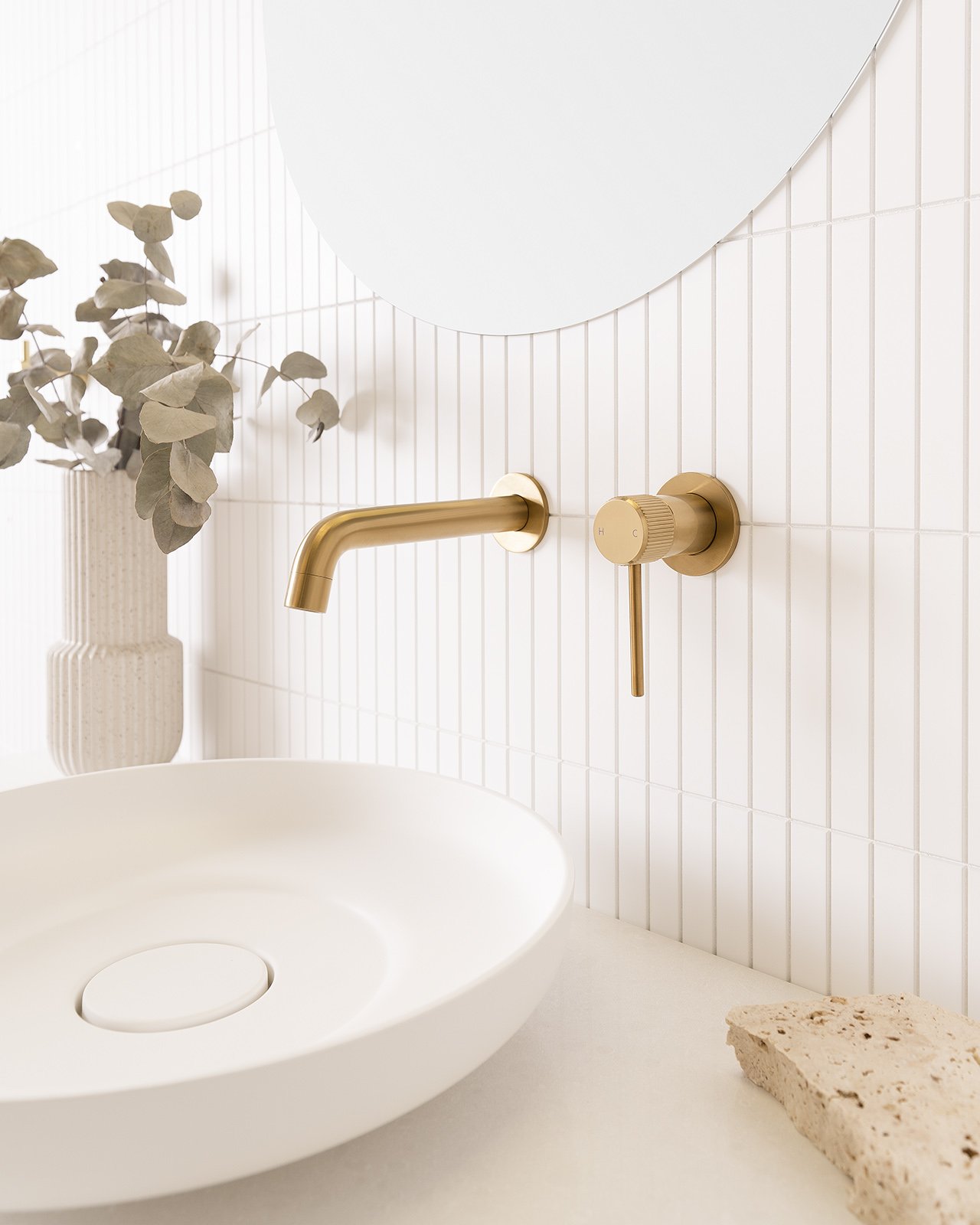 Modern bathroom sink with white round basin, gold faucet, gold handle, white tiled wall, vase with dried eucalyptus, mirror, and a piece of stone on the countertop.