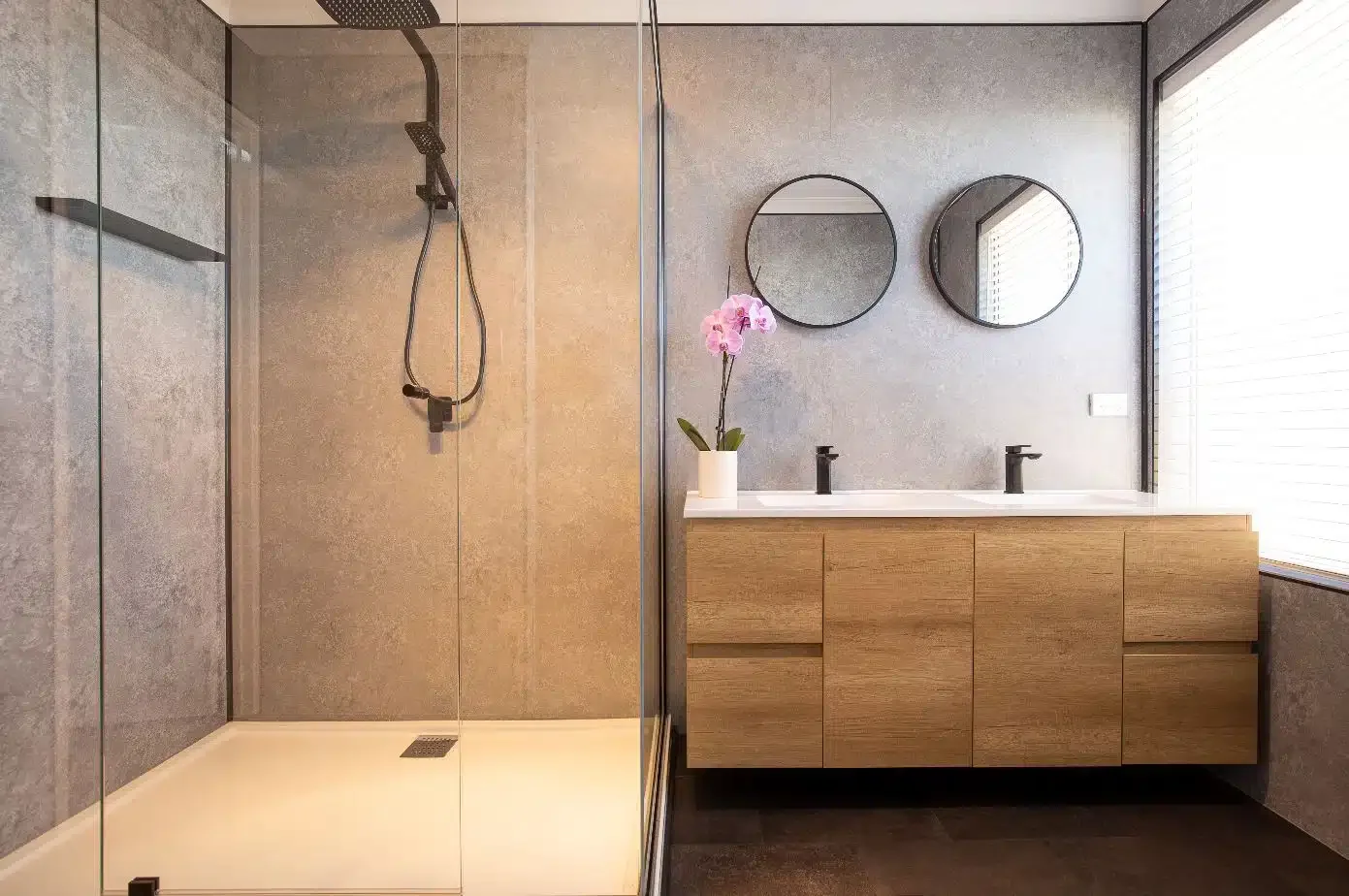 Modern bathroom with a walk-in shower, wooden vanity with two sinks, two round mirrors, and a vase of pink orchids on the countertop.