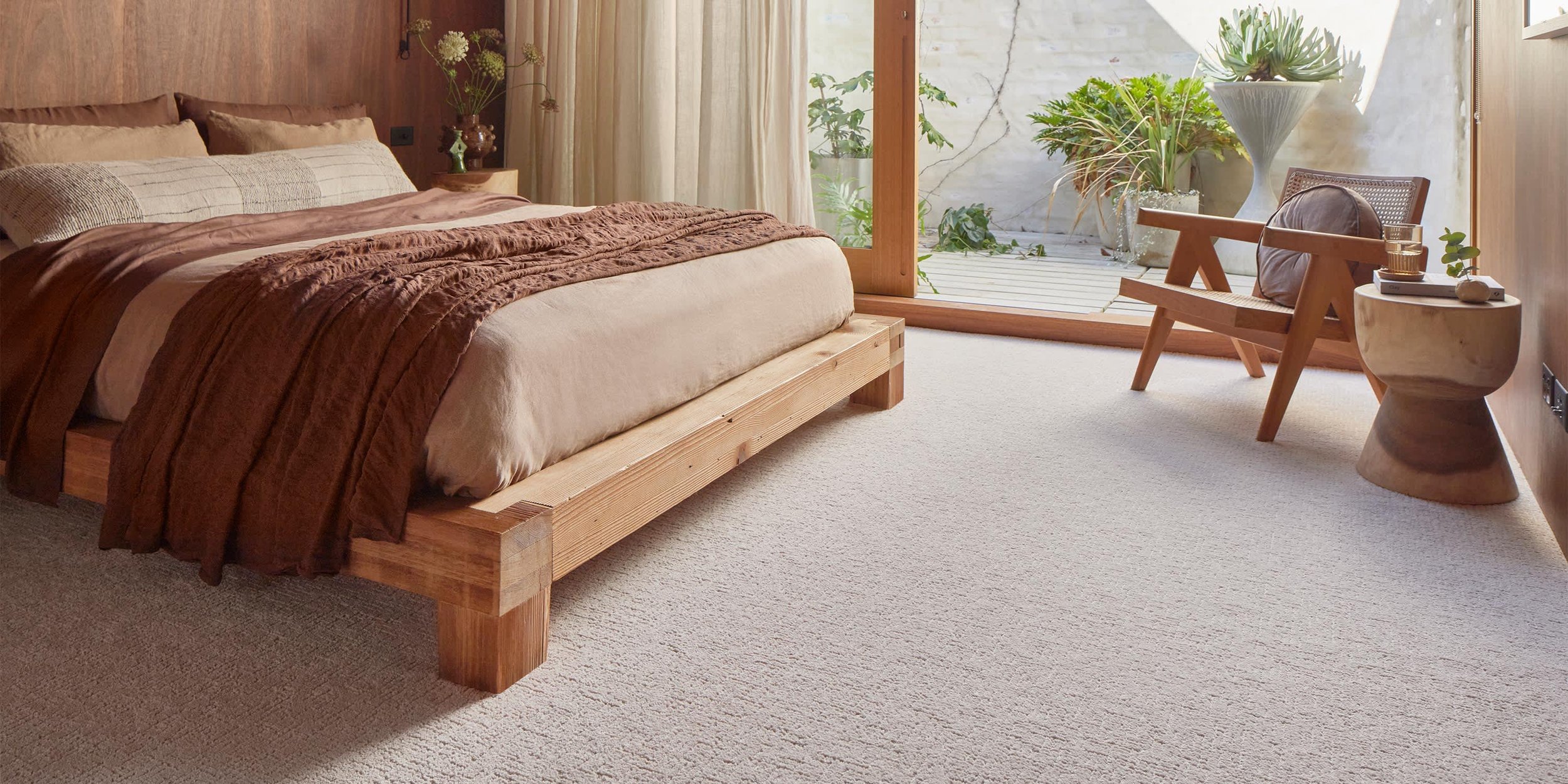 A bedroom with a wooden bed, brown bedding, and a side table with decorative objects. There is a glass door leading to a patio with plants and an outdoor seating area.