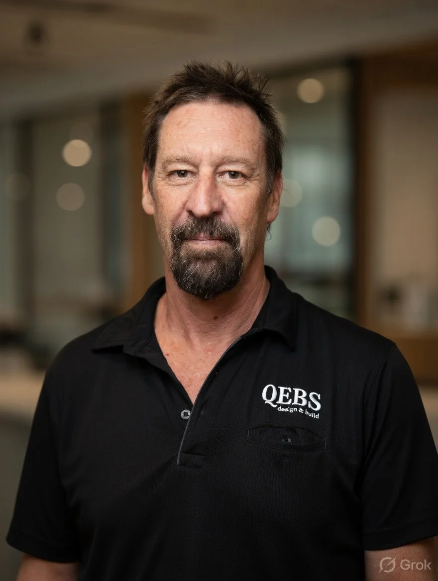 A middle-aged man with brown hair and a beard wearing a black polo shirt with 'QEB'S design & build' logo, posing indoors with a blurred background.