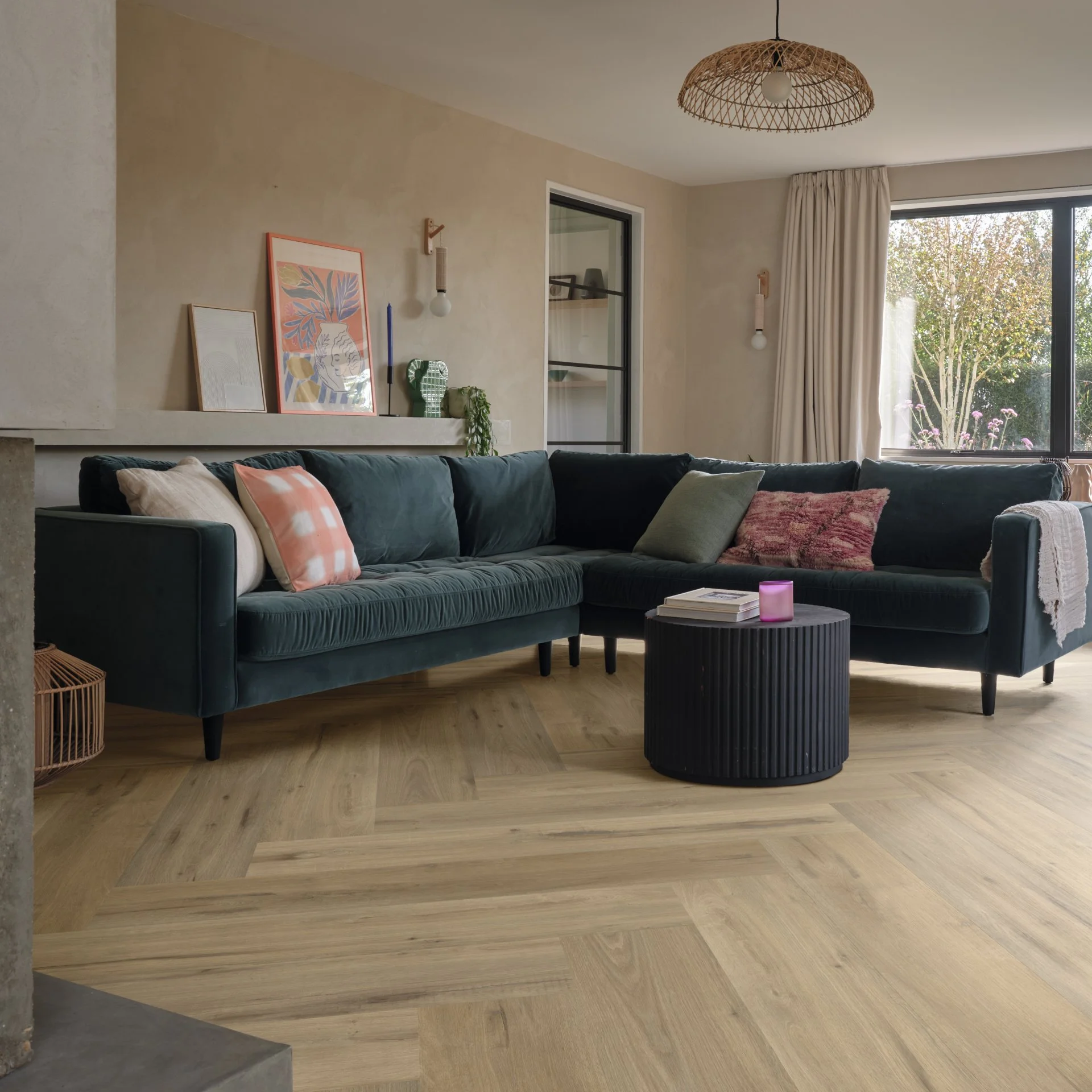 Living room with dark green sectional sofa, patterned throw pillows, black round coffee table, light wood flooring, large window with cream curtains, wall art, and modern lighting fixtures.