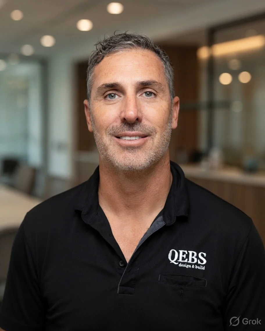 A middle-aged man with short gray hair and a beard, dressed in a black polo shirt with 'Q.E.B.S design & build' embroidered on it, smiling slightly in an office setting.