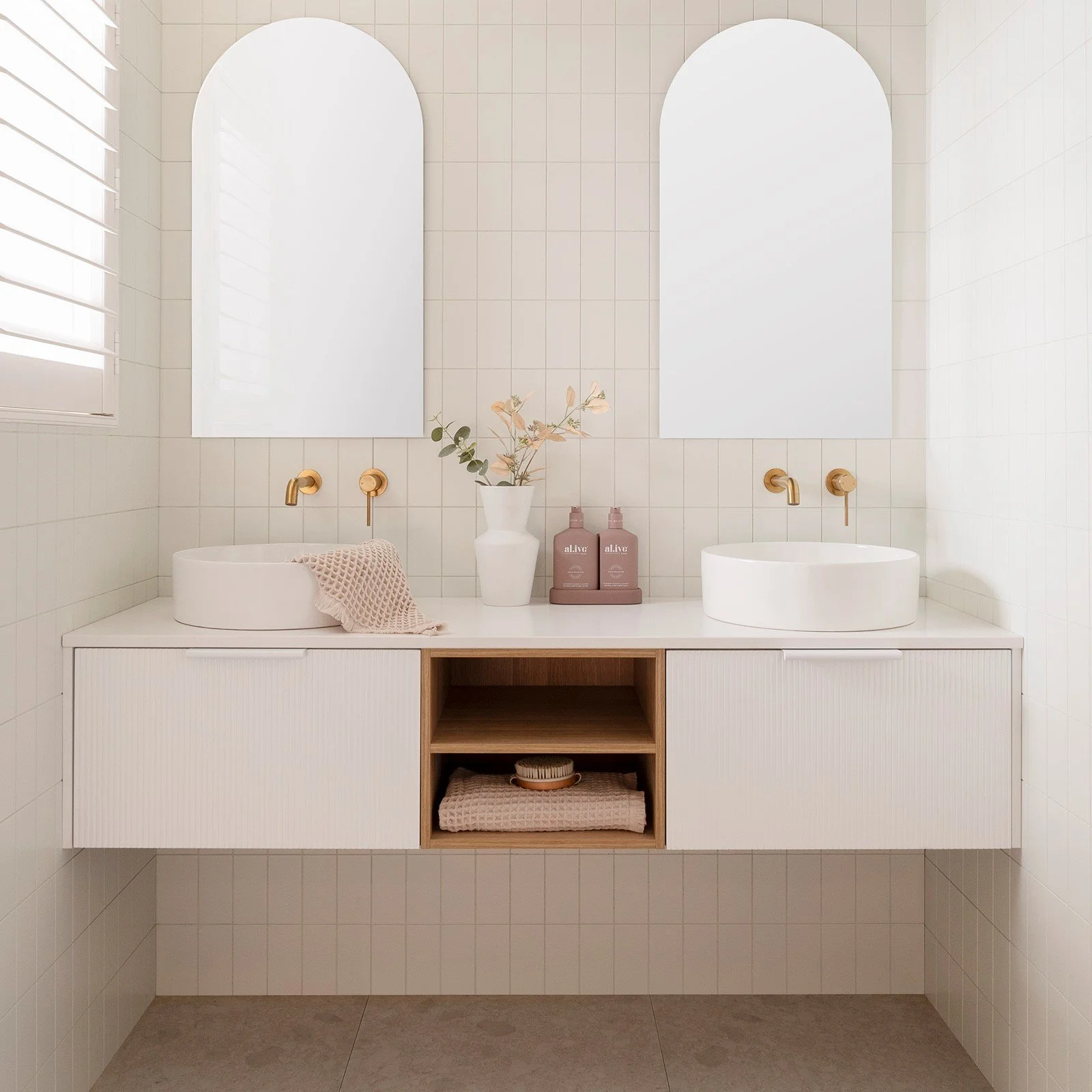 A modern bathroom with a double vanity, featuring two oval vessel sinks with gold faucets. There are two large arched mirrors above the sinks, and a window with white blinds on the left. The vanity has a white cabinet with an open shelf in the middle