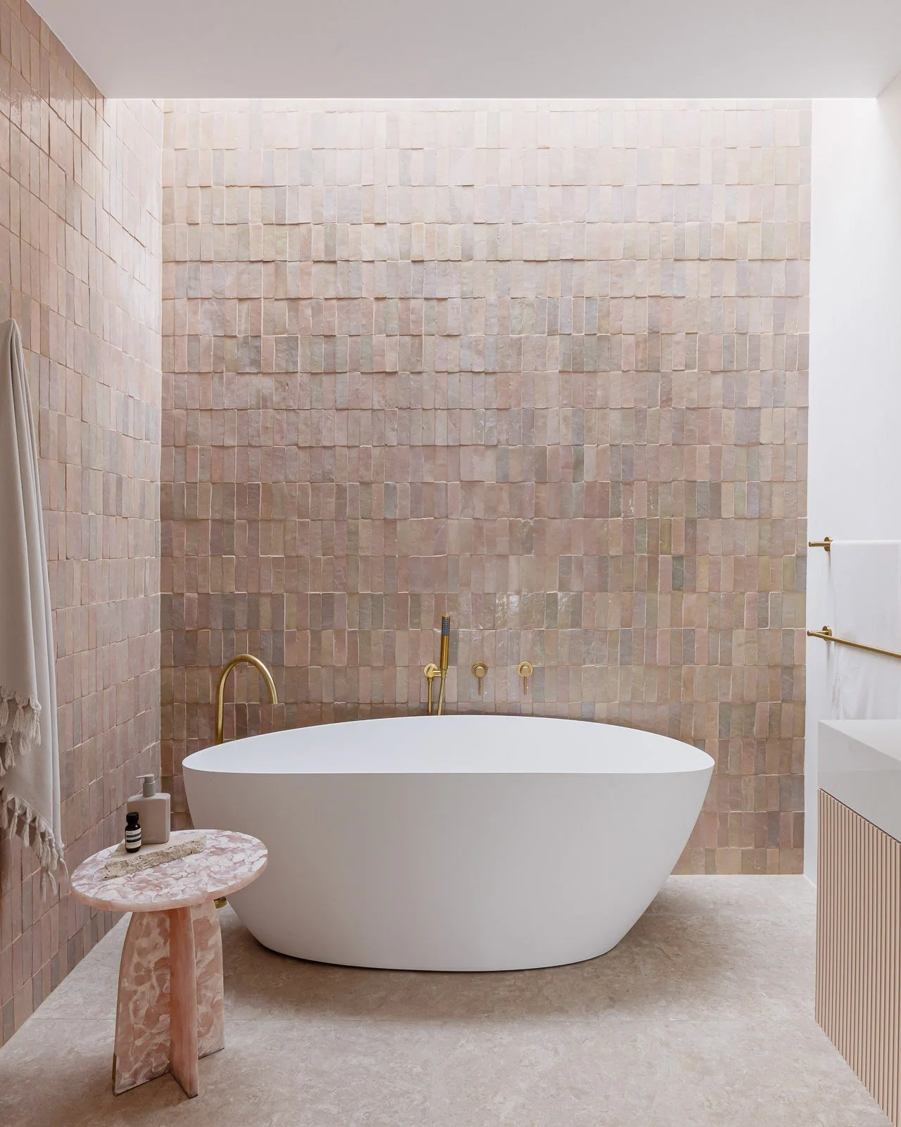 Modern bathroom with a pink tiled wall, freestanding white bathtub, gold fixtures, and a small pink marble side table with soap and lotion bottles.