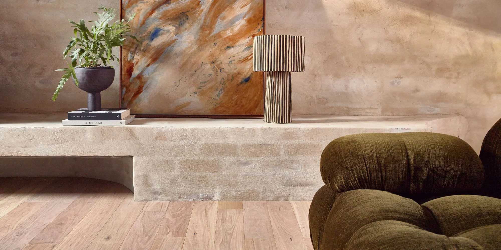 A beige stone or concrete ledge with a dark green upholstered armchair in the foreground. On the ledge, there is a dark vase with green plant, a stack of books, a large abstract art piece with warm tones, and a striped table lamp with a cylindrical shape. The background wall is made of beige stone or plaster.