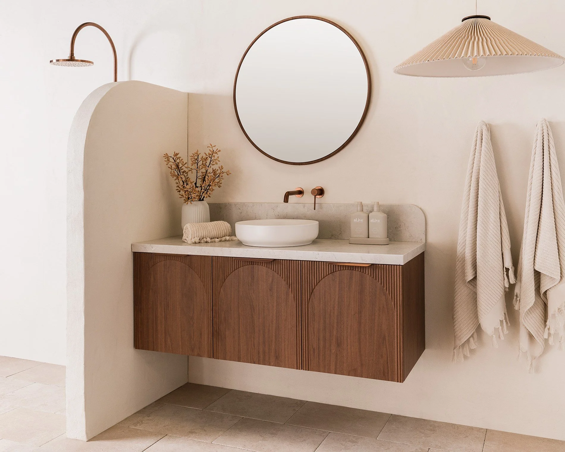 Modern bathroom vanity with a round vessel sink, large mirror, and wall-mounted faucet. Decor includes a vase with flowers, rolled towel, bottles of lotion or soap, and hanging towels. Features soft neutral tones and minimalist design.