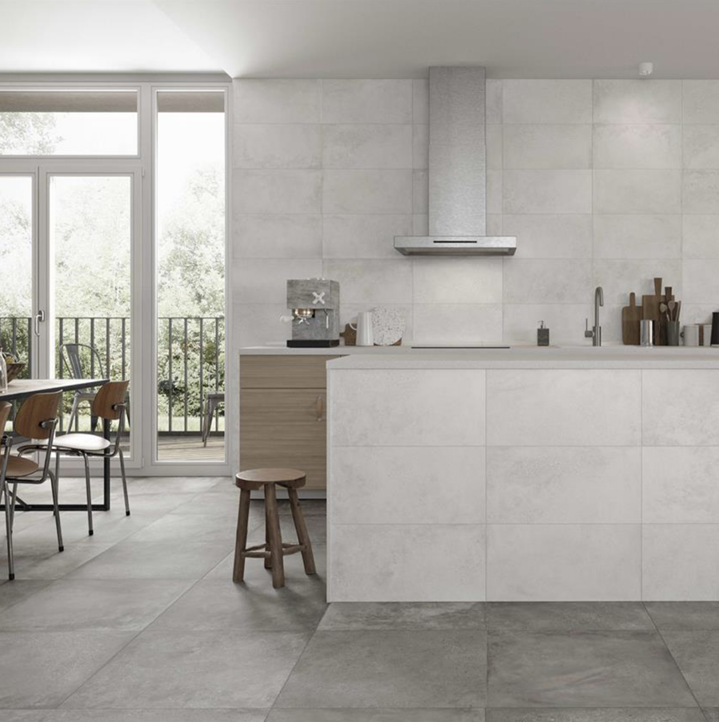 Modern kitchen with white tiled walls and gray floor, wooden chairs and table, a small wooden stool, large glass doors opening to a balcony with trees outside, and a sleek stainless steel range hood over the countertop.