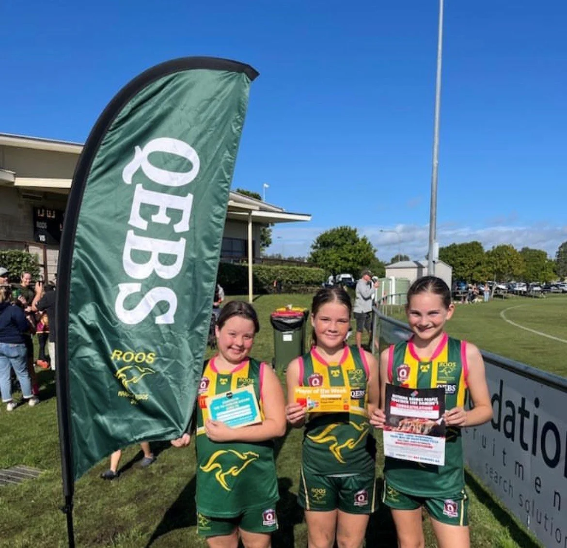 Three young girls in sports uniforms holding flyers and medals are standing outdoors near a green feather flag and a sports field, celebrating at a sporting event.