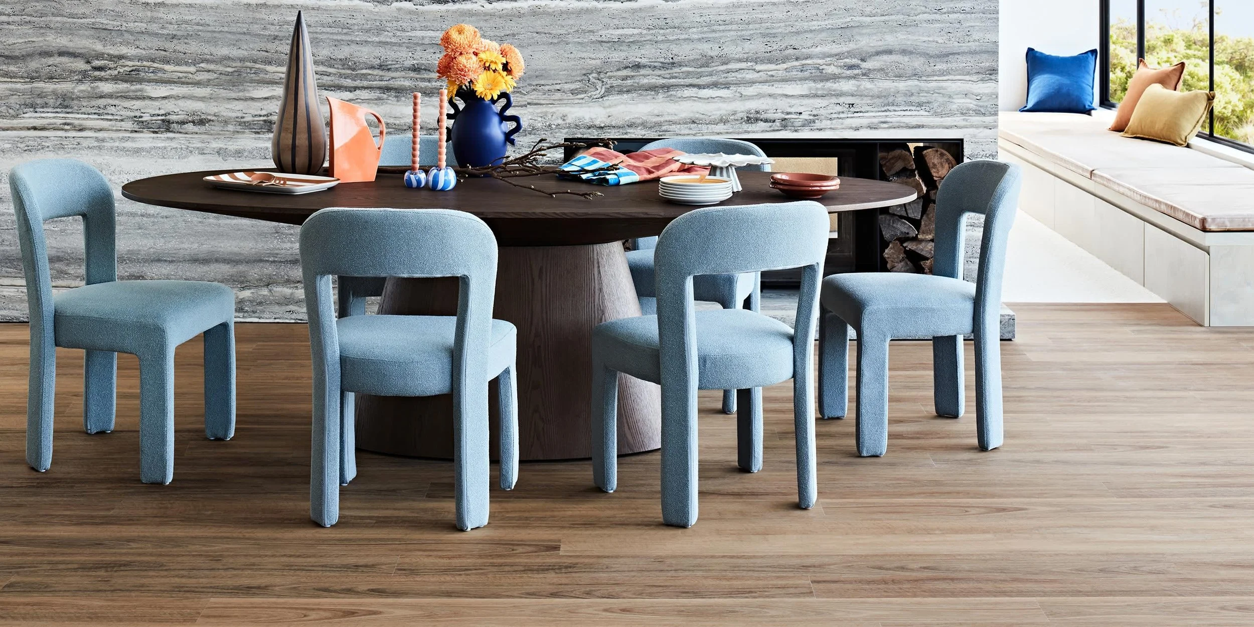 A dining room with a round dark wood table surrounded by six light blue upholstered chairs. The table has various decorative items, including a tall striped vase, a blue pitcher, two pink candles, a branch, and some plates. In the background, there i