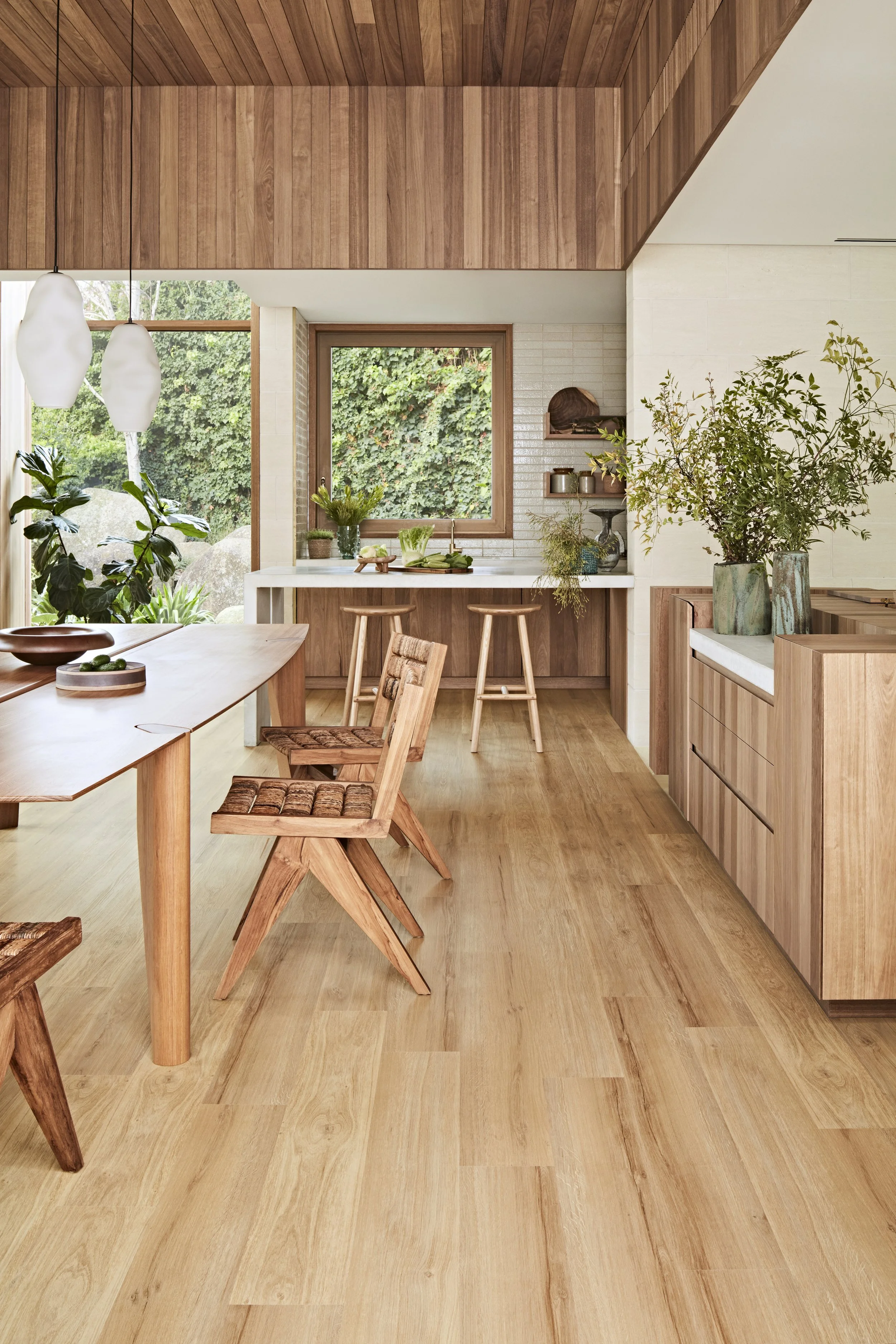 A cozy, modern kitchen with wood-paneled ceiling, wood flooring, white walls, a window overlooking greenery, a marble countertop, potted plants, wooden chairs, and decorative shelves.
