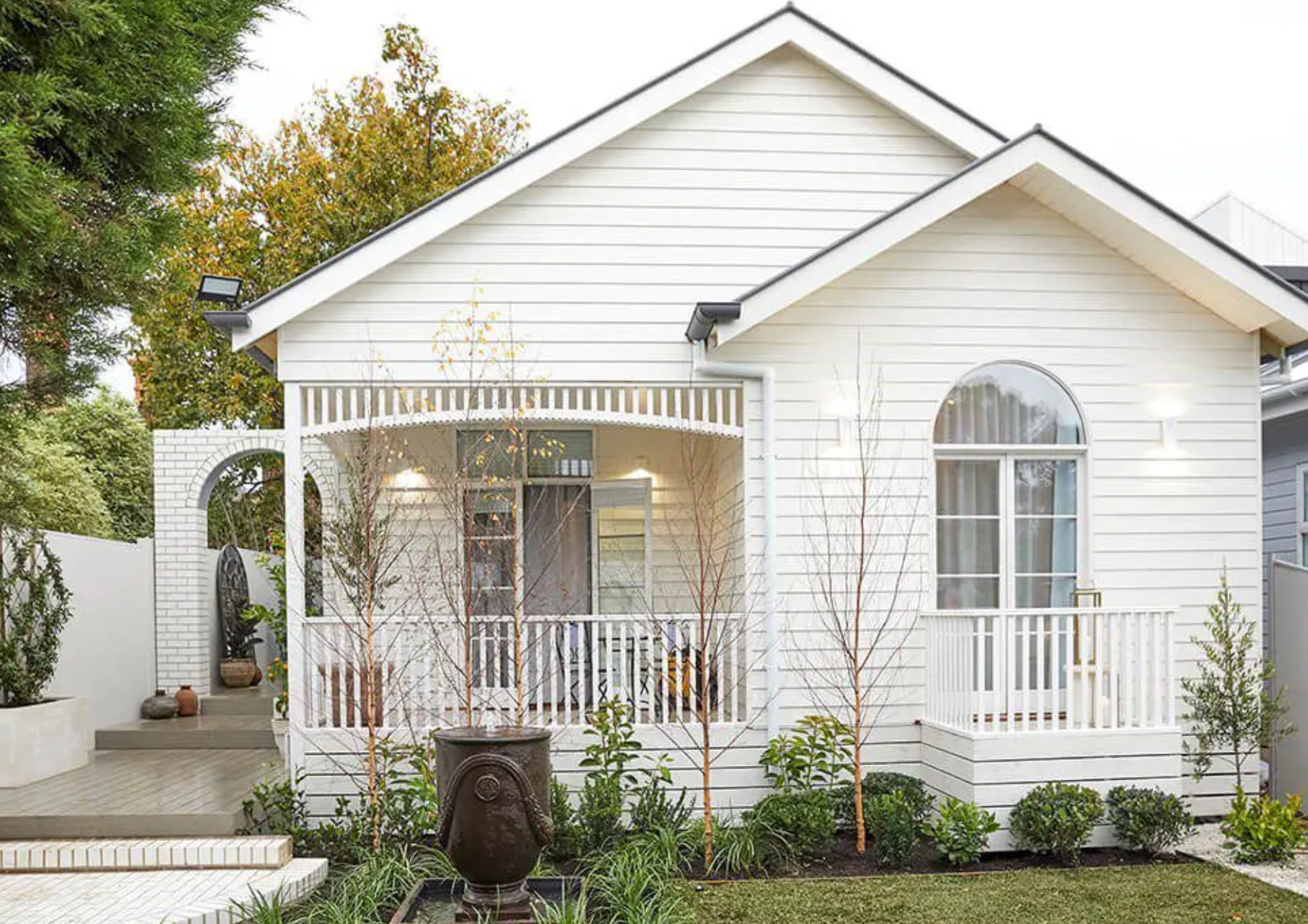 Front view of a white house with a porch, arched window, and landscaping with small trees and bushes.