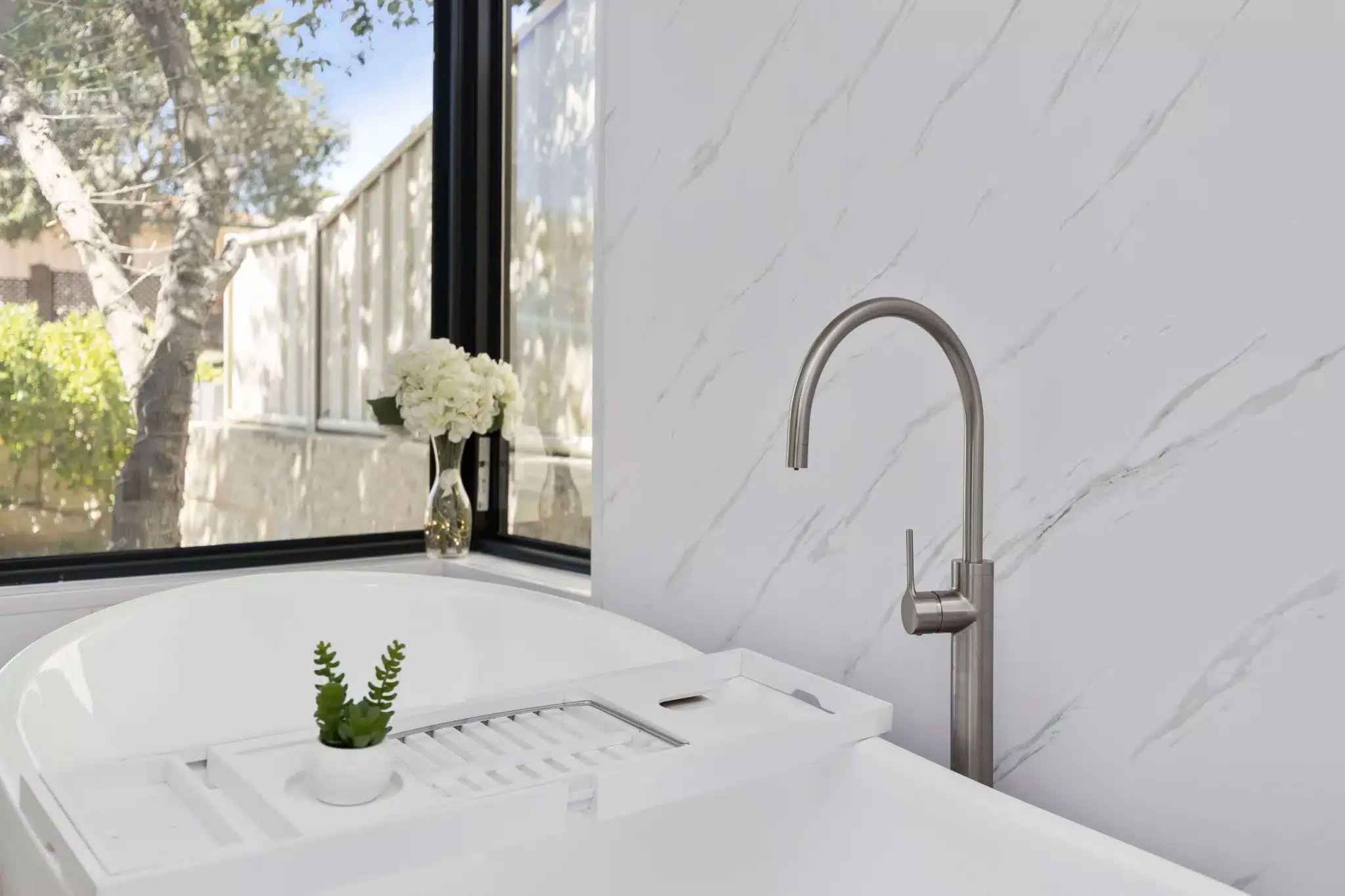 Bathroom with white marble wall, modern chrome faucet, bathtub with a built-in rack holding a small potted succulent plant, and a window with a vase of white flowers outside.