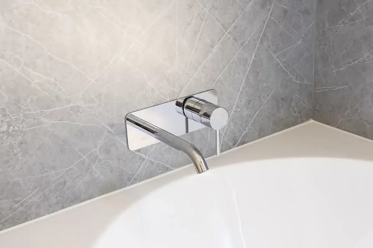 Close-up of a modern wall-mounted bathroom faucet with a chrome finish, attached to a gray marble wall over a white bathtub.