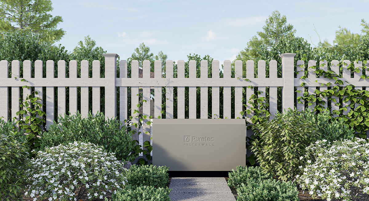 A white wooden fence with greenery and vines growing on its sides, a landscaped garden with bushes and flowering plants in the foreground, and a cloudy sky in the background.