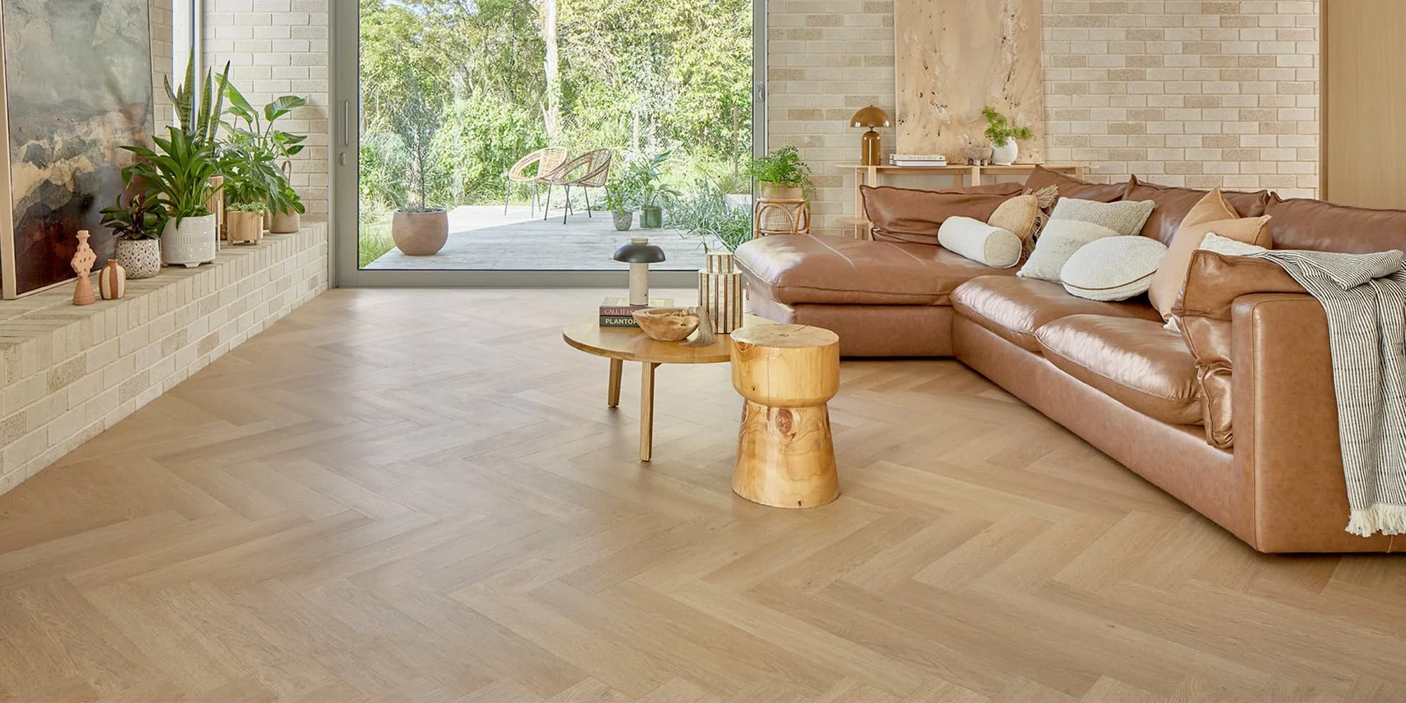 A living room with a brown leather sectional sofa, a round wooden coffee table, and a sliding glass door leading to an outdoor patio with greenery.