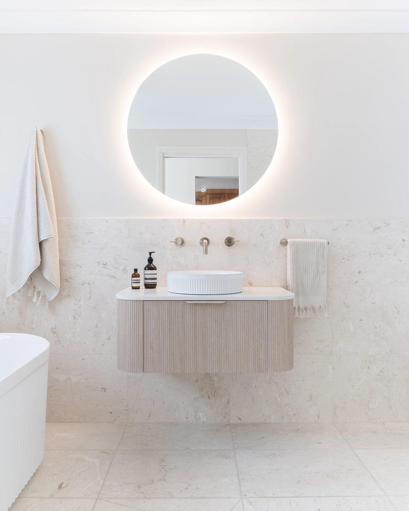 Modern bathroom with a round backlit mirror above a white and beige floating vanity, a vessel sink, soap dispenser, and hand towel, with beige tiled walls and floors.
