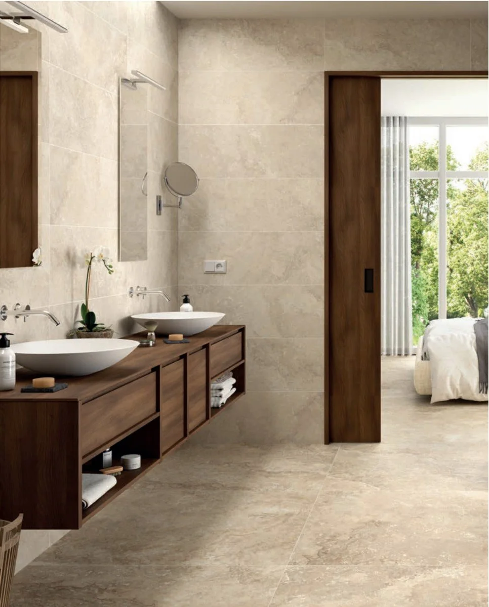 Modern bathroom with beige tile walls and floor, wooden vanity with two vessel sinks, and wall-mounted mirrors. An open doorway reveals a bedroom with large windows and view of green trees.