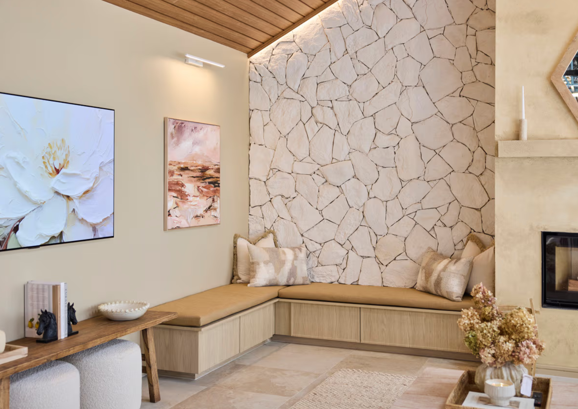 A cozy living room corner with beige upholstery, decorative pillows, a wood-paneled ceiling, stone accent wall, wall art, and a wooden coffee table with flowers and candles.