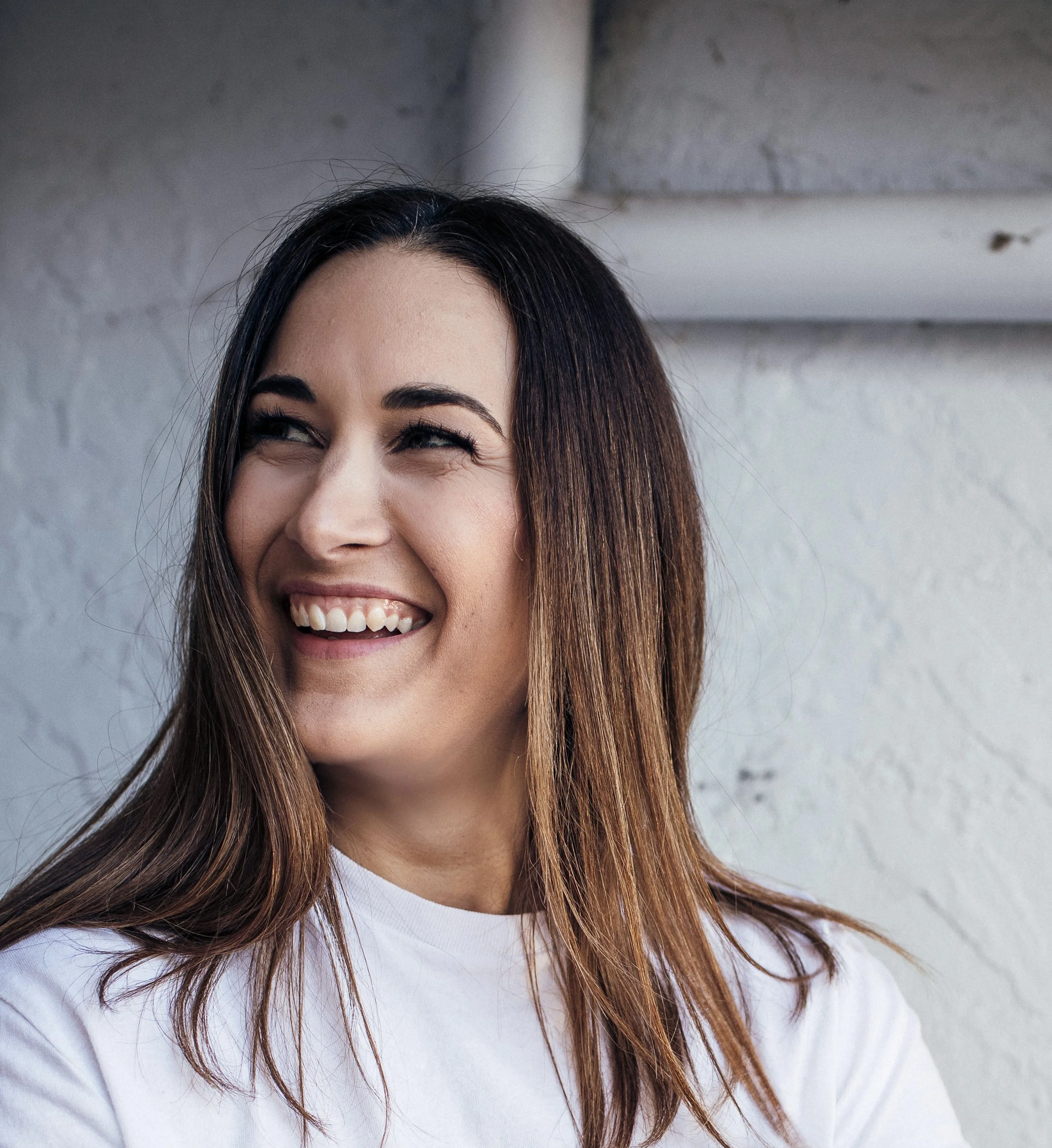 A woman with long brown hair smiling and looking to the side, standing against a white brick wall.