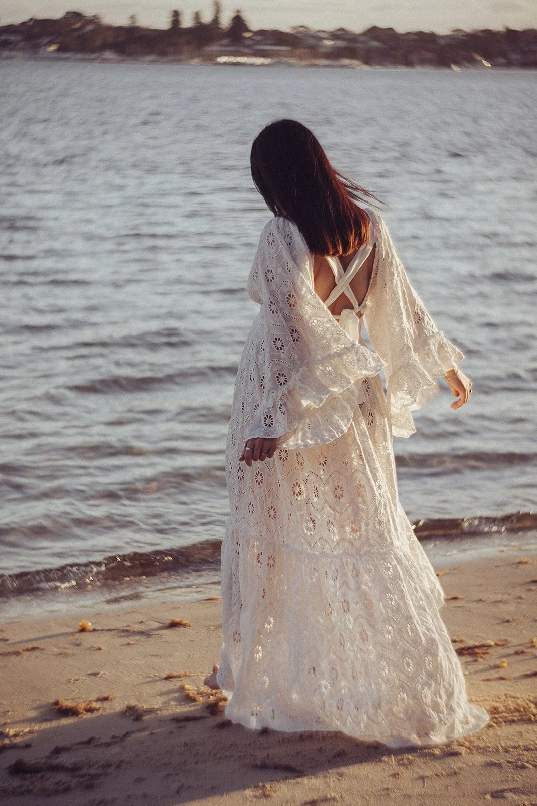 Woman in a long, white lace dress standing on the beach near the water, facing away from the camera, with her head slightly bowed.