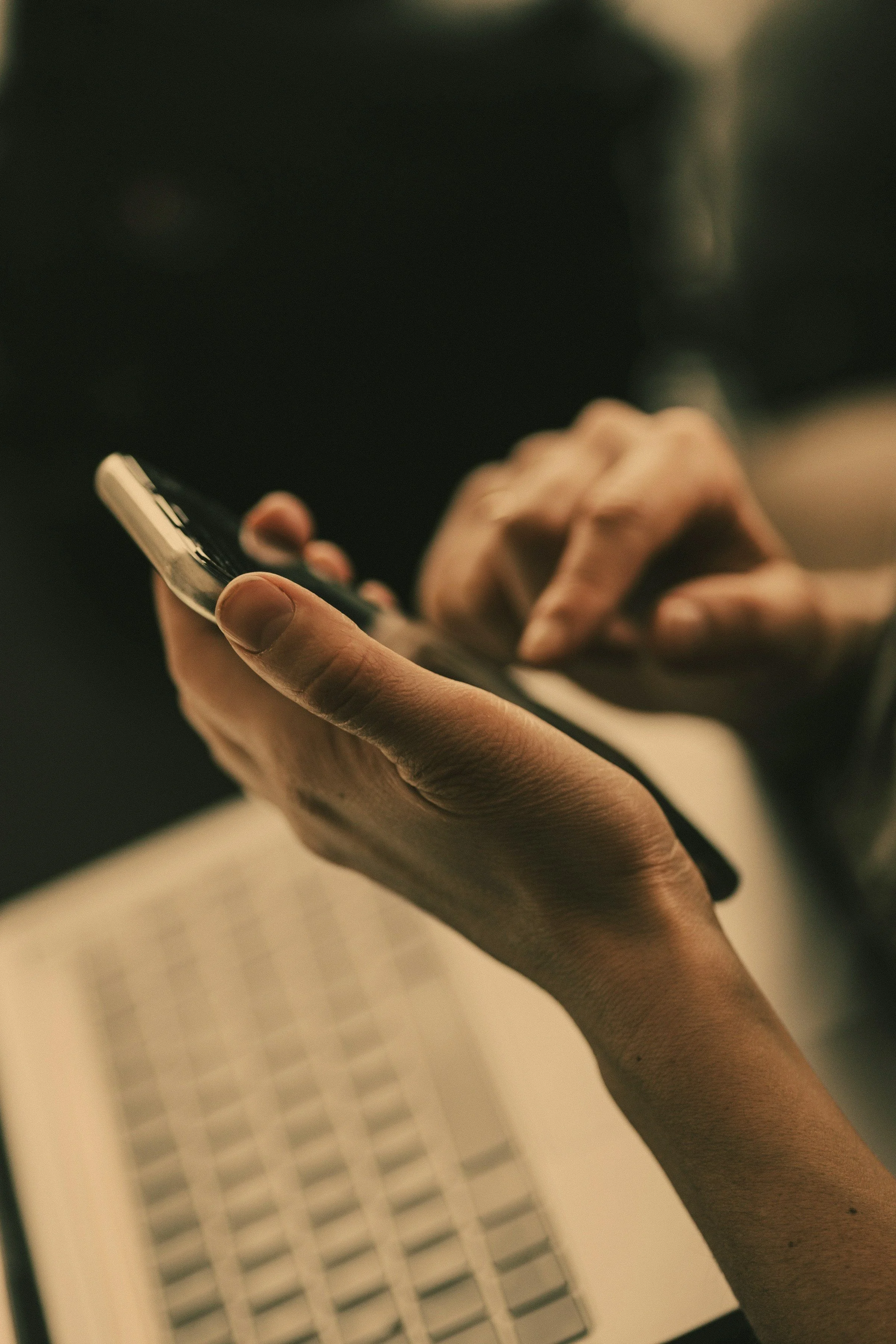 Close-up of a person's hand holding a smartphone, with a laptop keyboard visible in the background.
