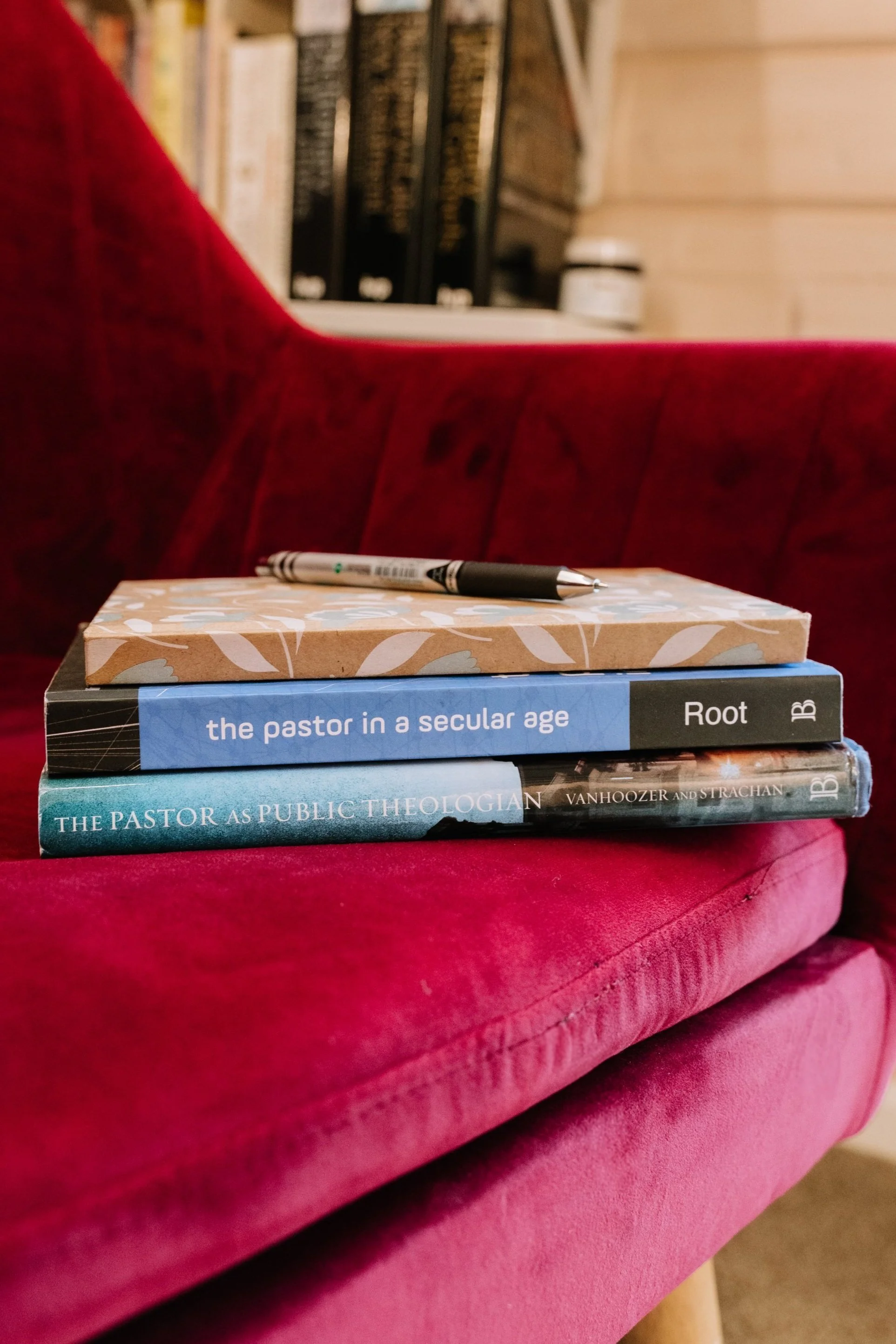Three books stacked on a red velvet sofa with a black and white pen resting on top.