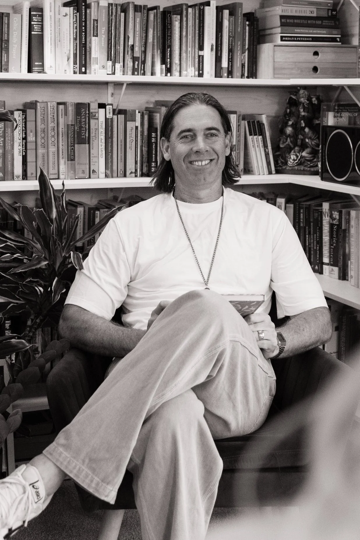 A man sitting in a chair, smiling, in front of a bookshelf filled with books, with a plant to his side.