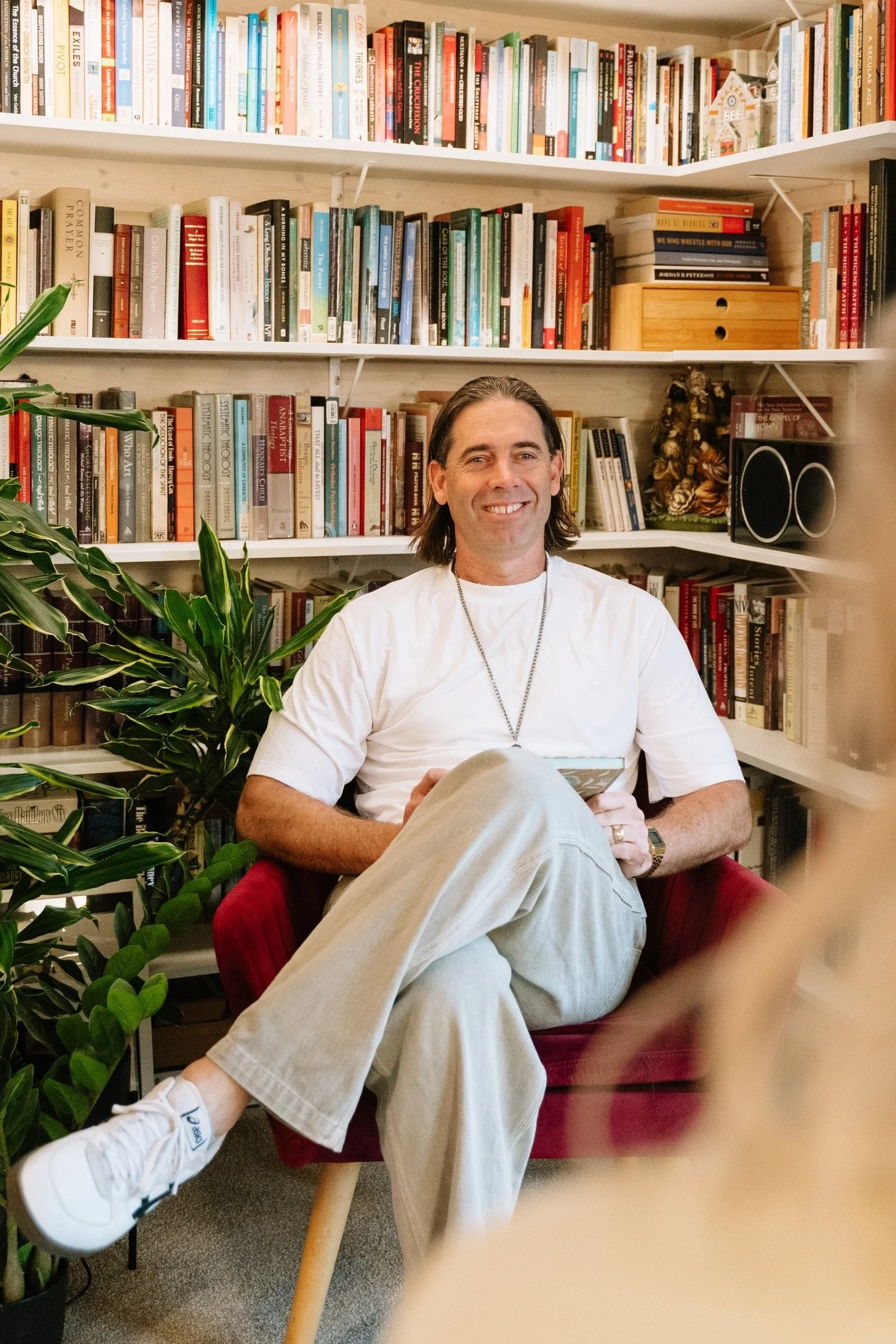 A man with long dark hair, wearing a white t-shirt and beige pants, is sitting cross-legged on a red chair in front of a bookshelf filled with various books. He is smiling and holding a book in his right hand.