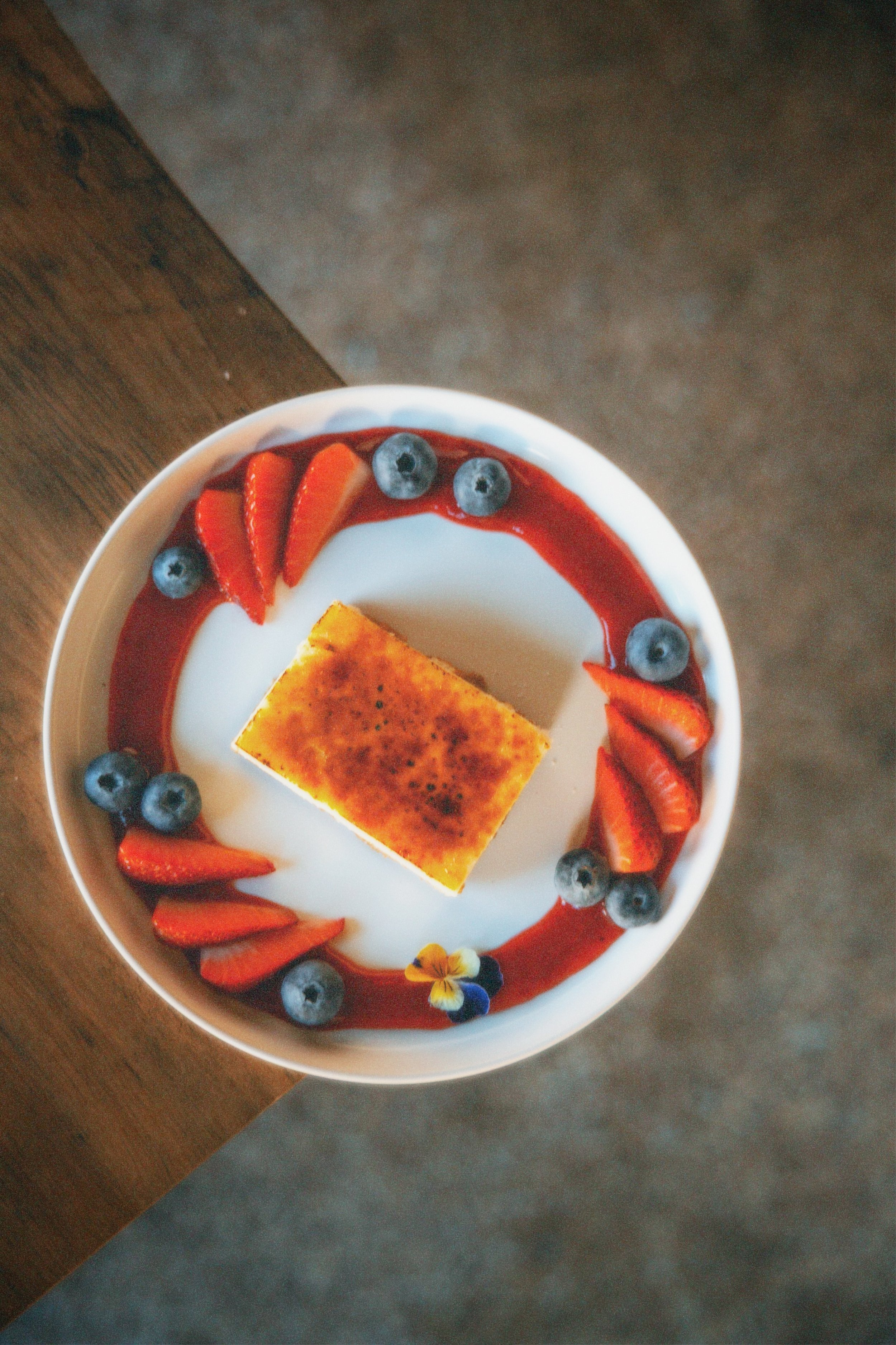 A dessert bowl with a square piece of crème brûlée surrounded by sliced strawberries, blueberries, red sauce, and edible flowers.