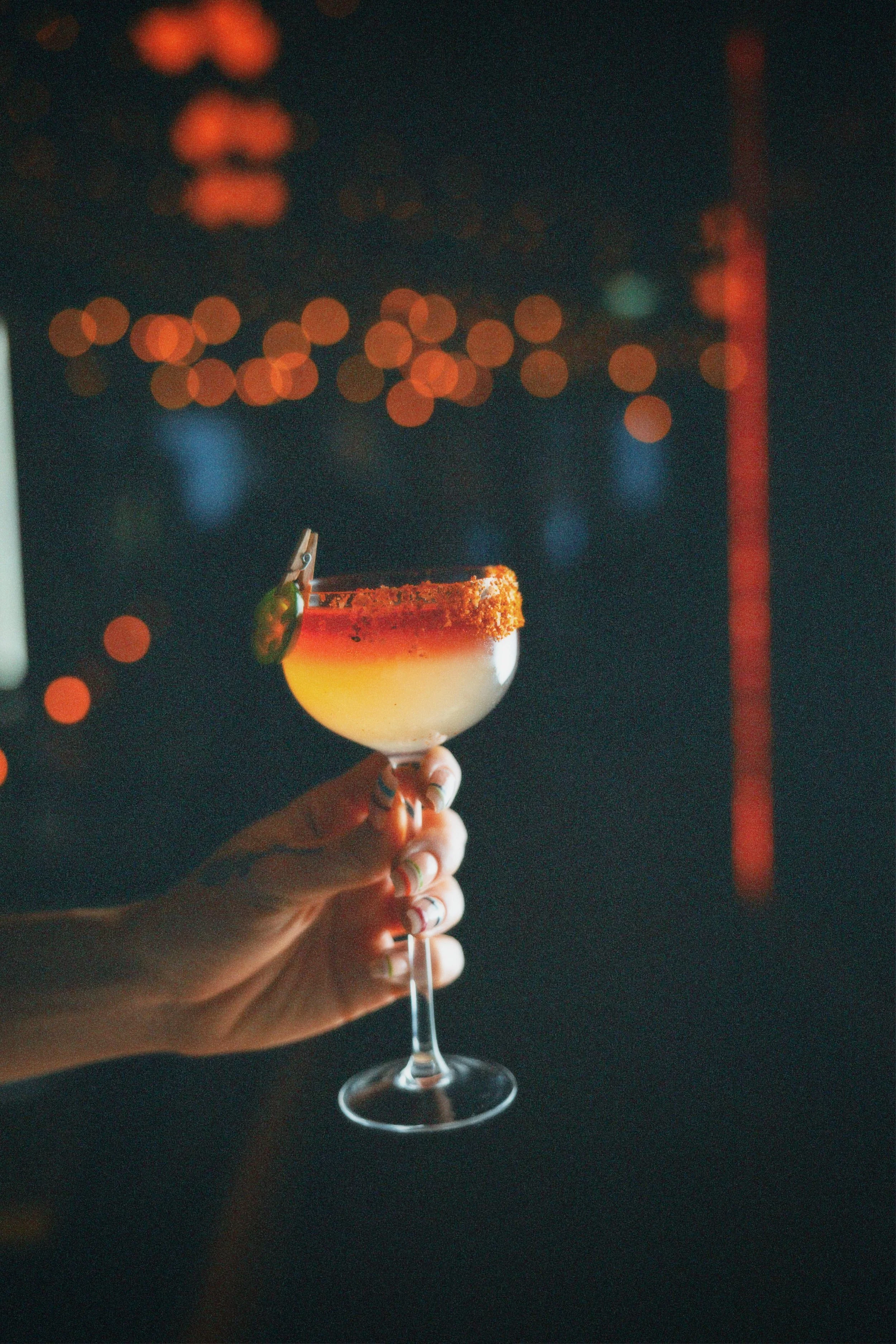 A person's hand holding a cocktail glass with a salted rim and a lime garnish, against a blurred background with orange bokeh lights.