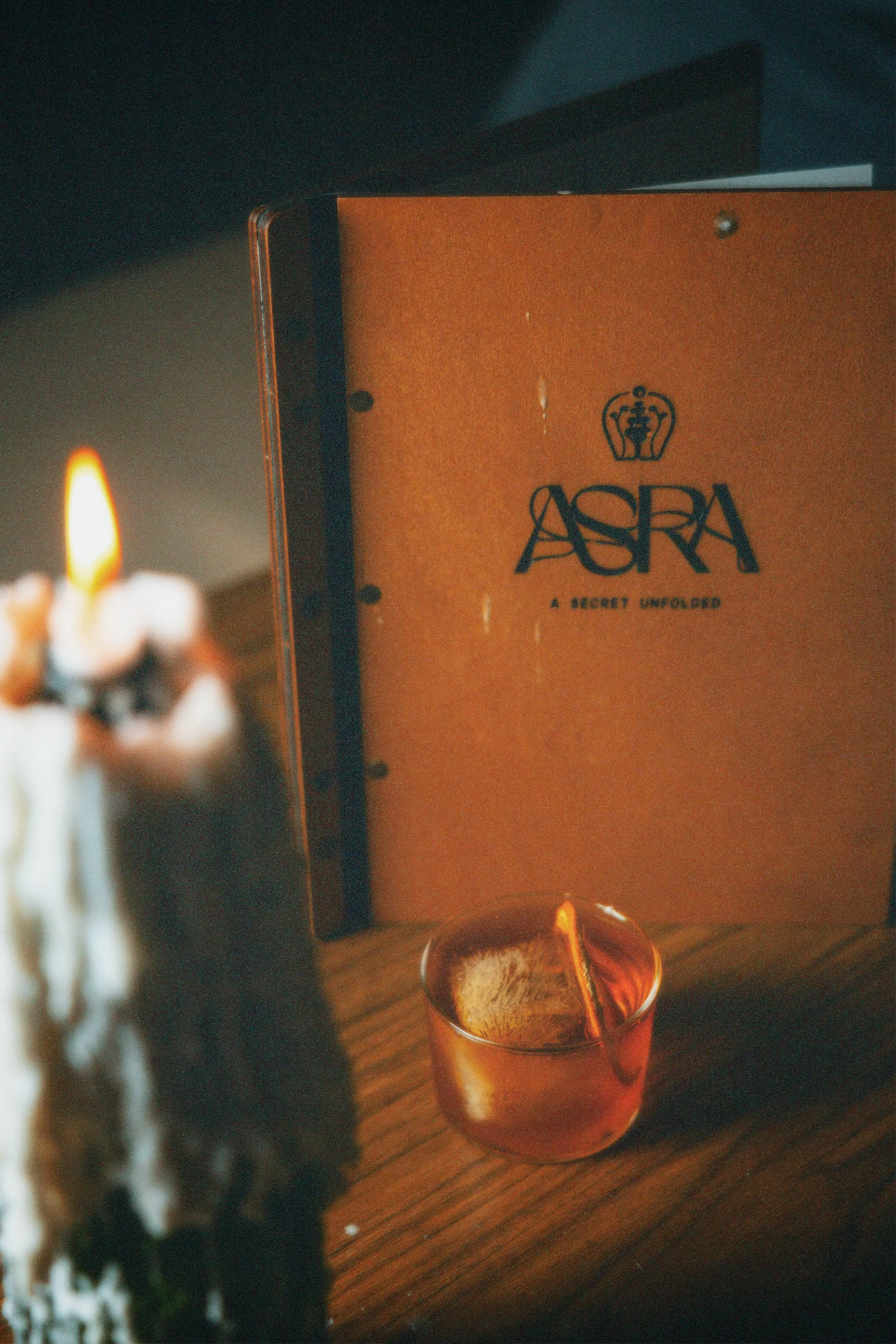 A menu titled 'ASARA: A Secret Unfolded' on a wooden table, with a burning candle in the foreground.