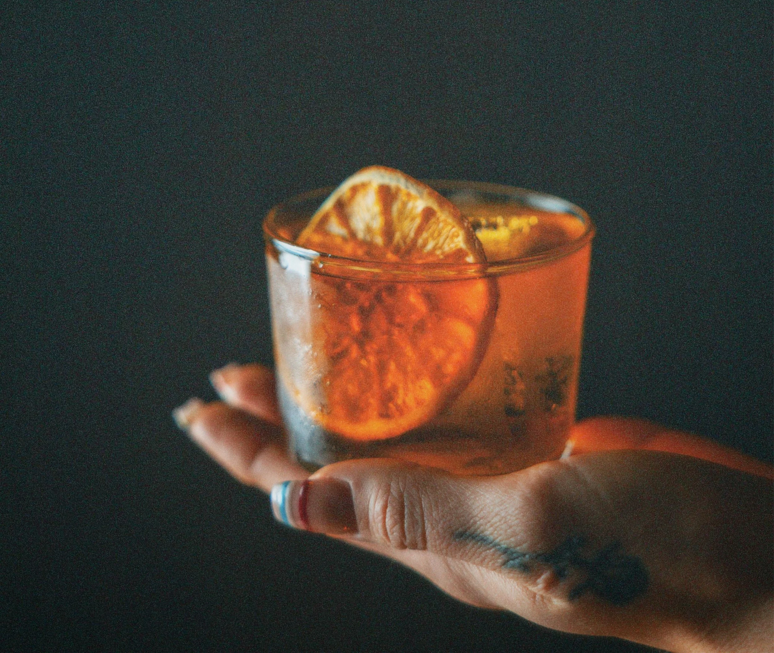 A hand holding a glass of orange-colored beverage with orange slices inside, against a dark background.
