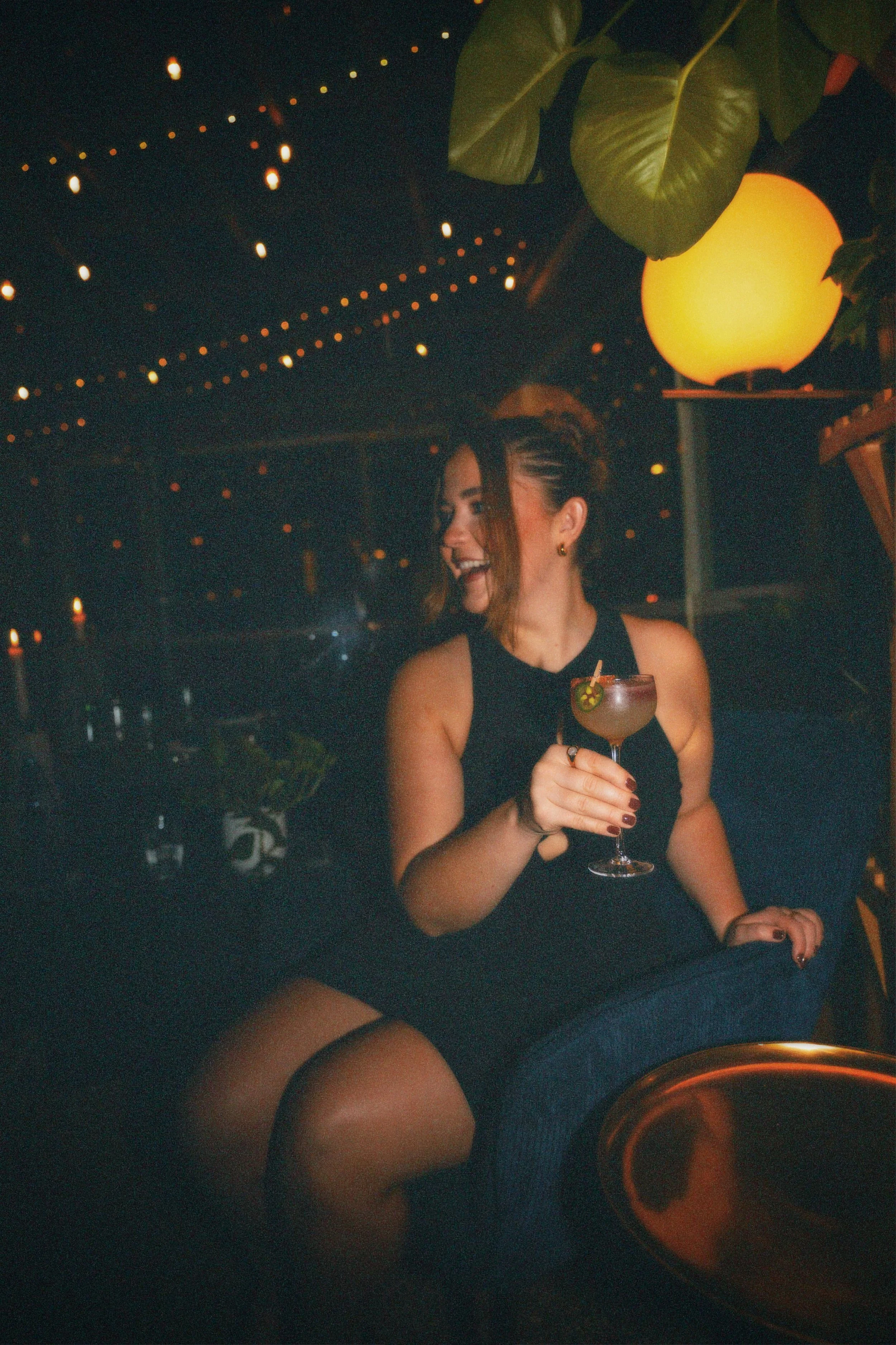 A woman in a black dress sitting in a dimly lit bar or lounge, holding a cocktail glass with a lime slice garnish, smiling and laughing. The background has string lights and large green leaves, with warm ambient lighting.