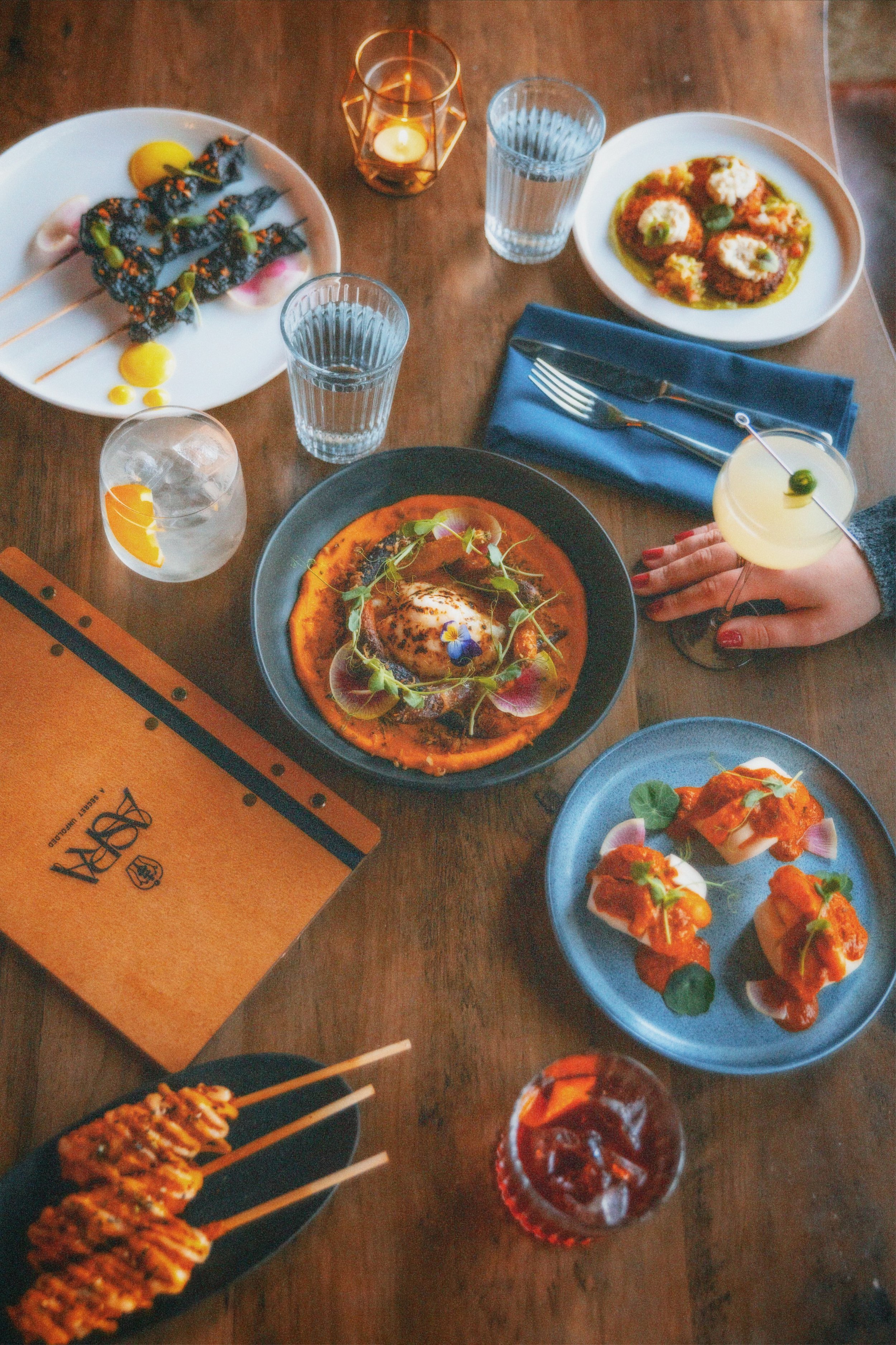A table with various dishes and drinks at a restaurant, including skewered meat, a savory dish with a poached egg, and a plate with wrapped appetizers, accompanied by water, cocktails, and a lit candle.