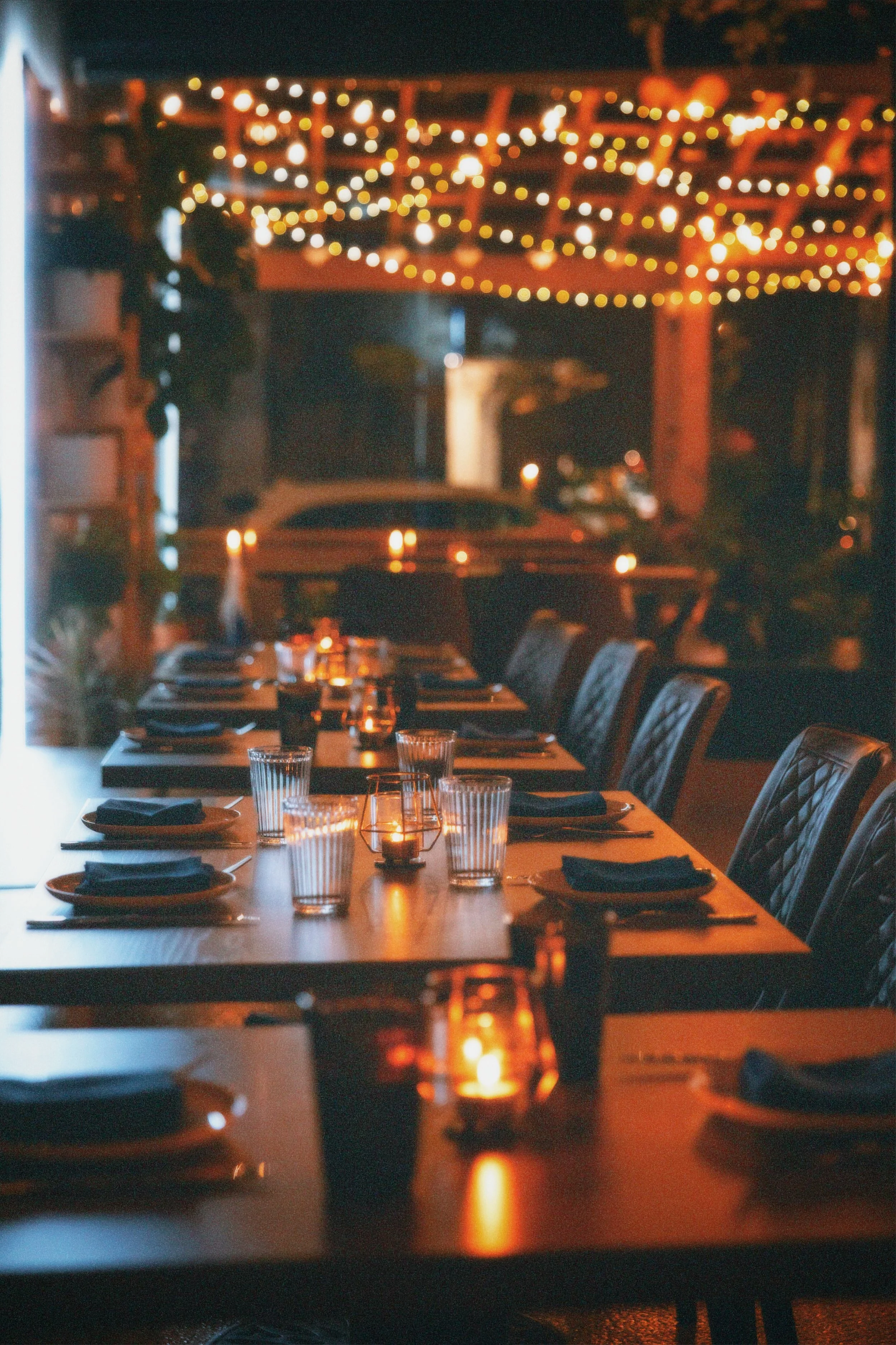 A dimly lit restaurant table set for dinner with glasses, plates, and utensils. String lights and lanterns create a warm, cozy atmosphere.