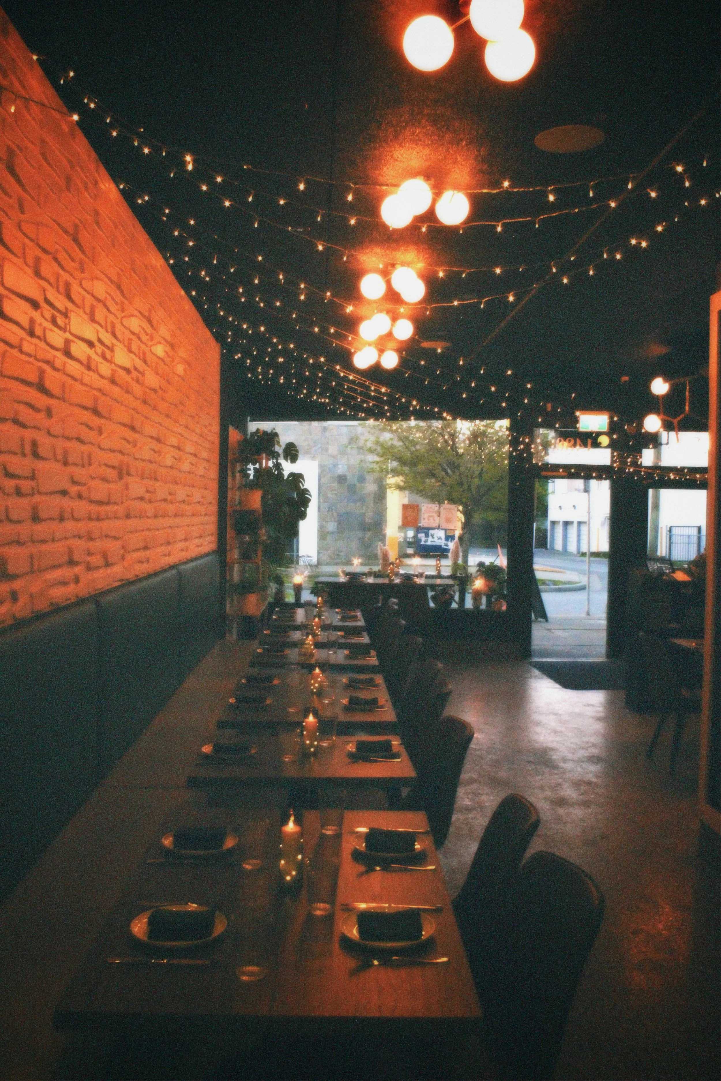 Indoor restaurant with a long dining table set with plates, glasses, napkins, and candles, illuminated by warm hanging lights and string lights, with an open view to the street outside and some greenery visible through the entrance.