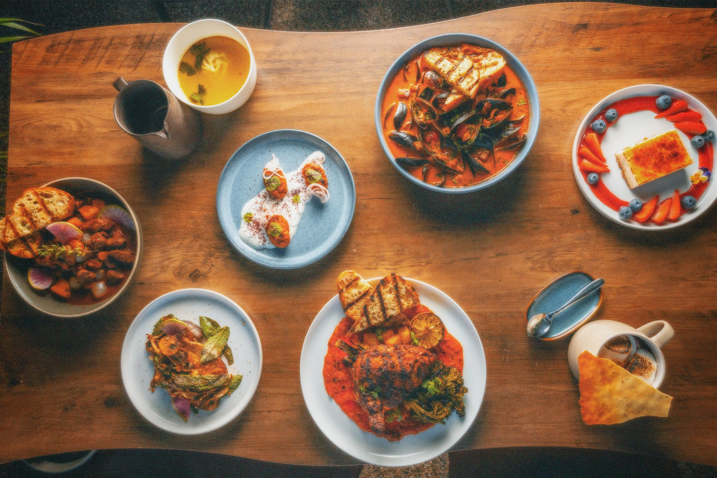 A wooden table with various plates of Indian cuisine, including curries, naan bread, rice, dessert, tea, and a coffee, arranged for a meal.