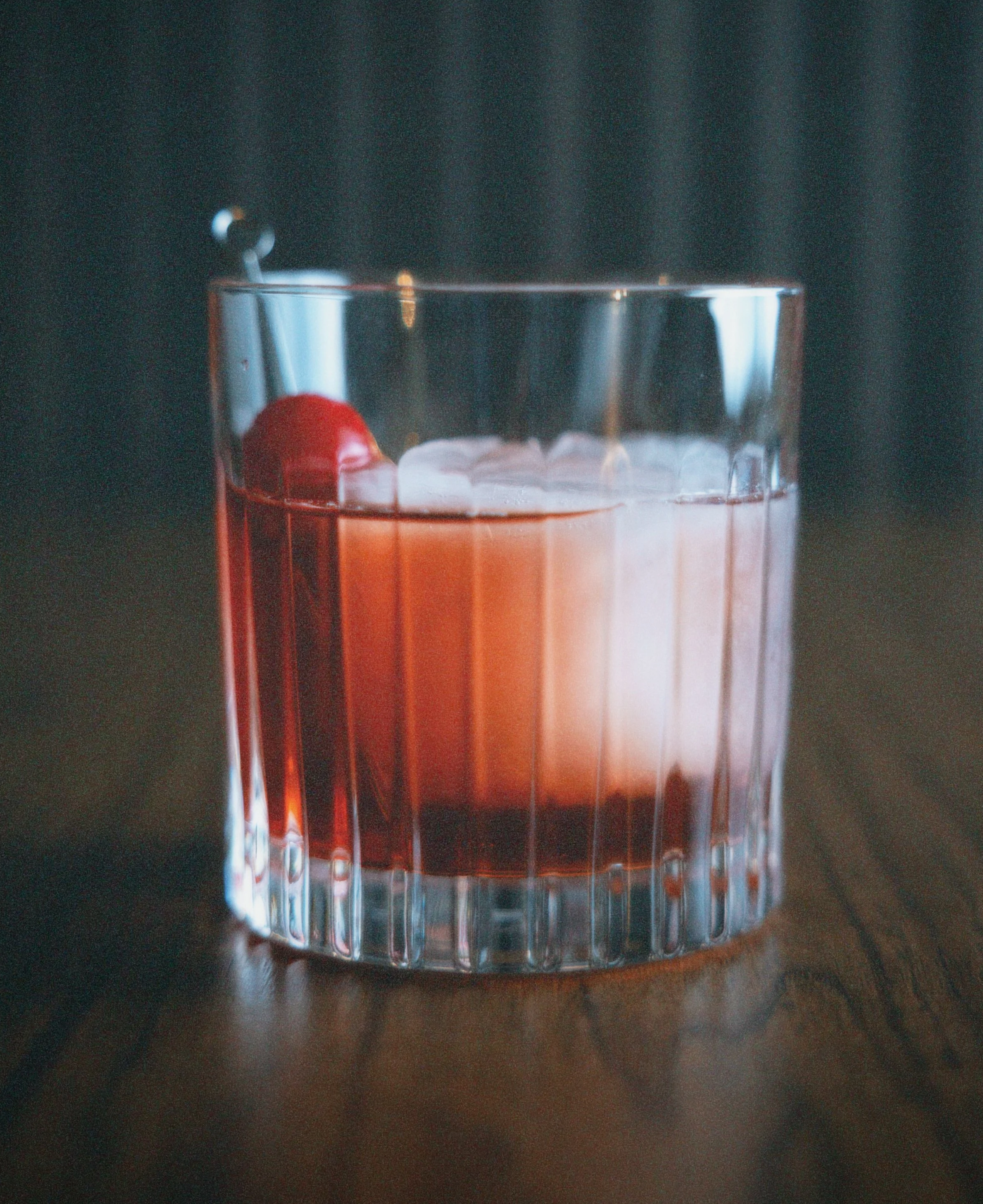 A glass of a pink cocktail with a cherry garnish and ice cubes on a wooden surface.