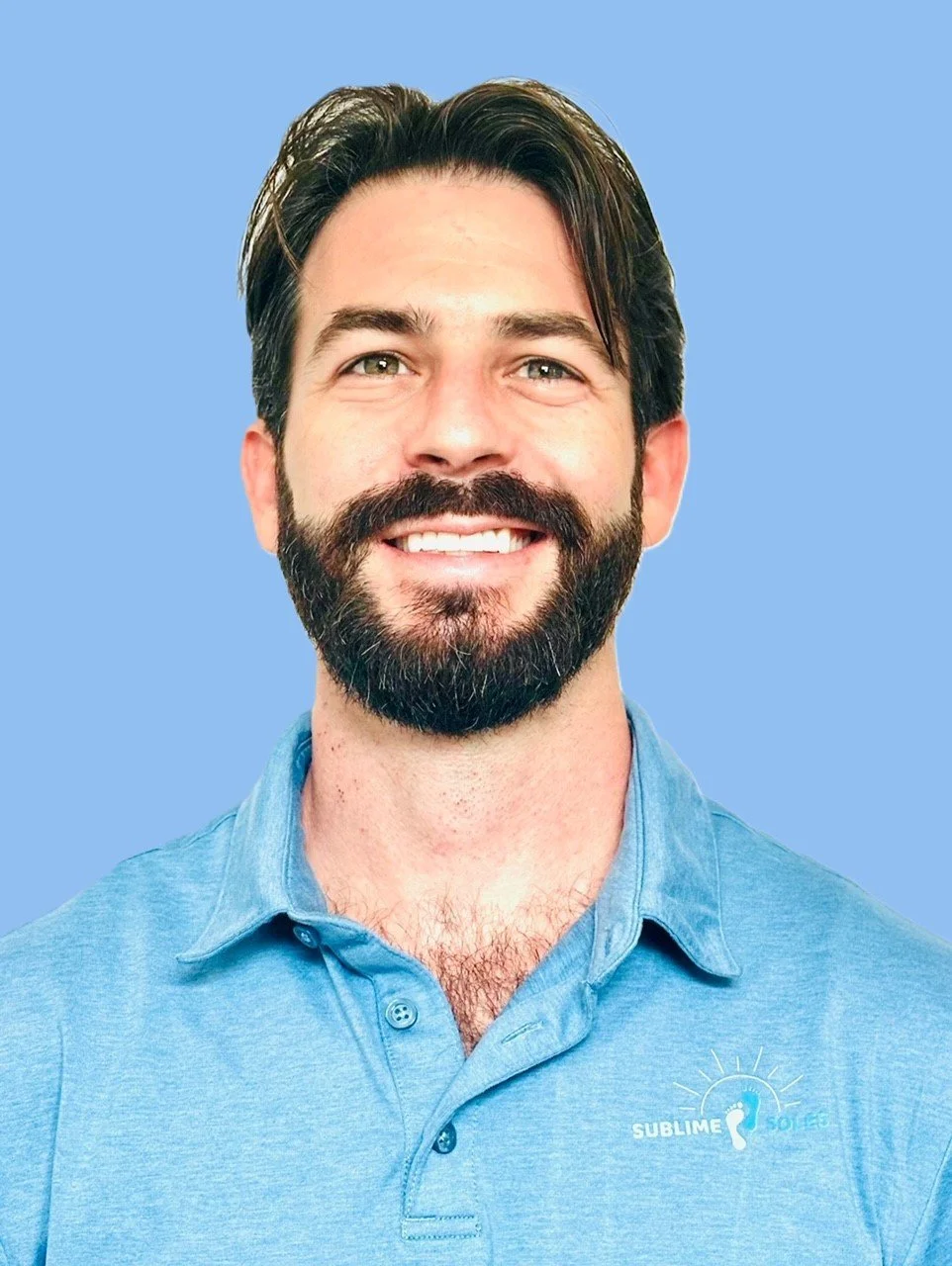 A smiling man with dark hair and a beard wearing a light blue polo shirt with a Sublime Soles logo against a blue background.