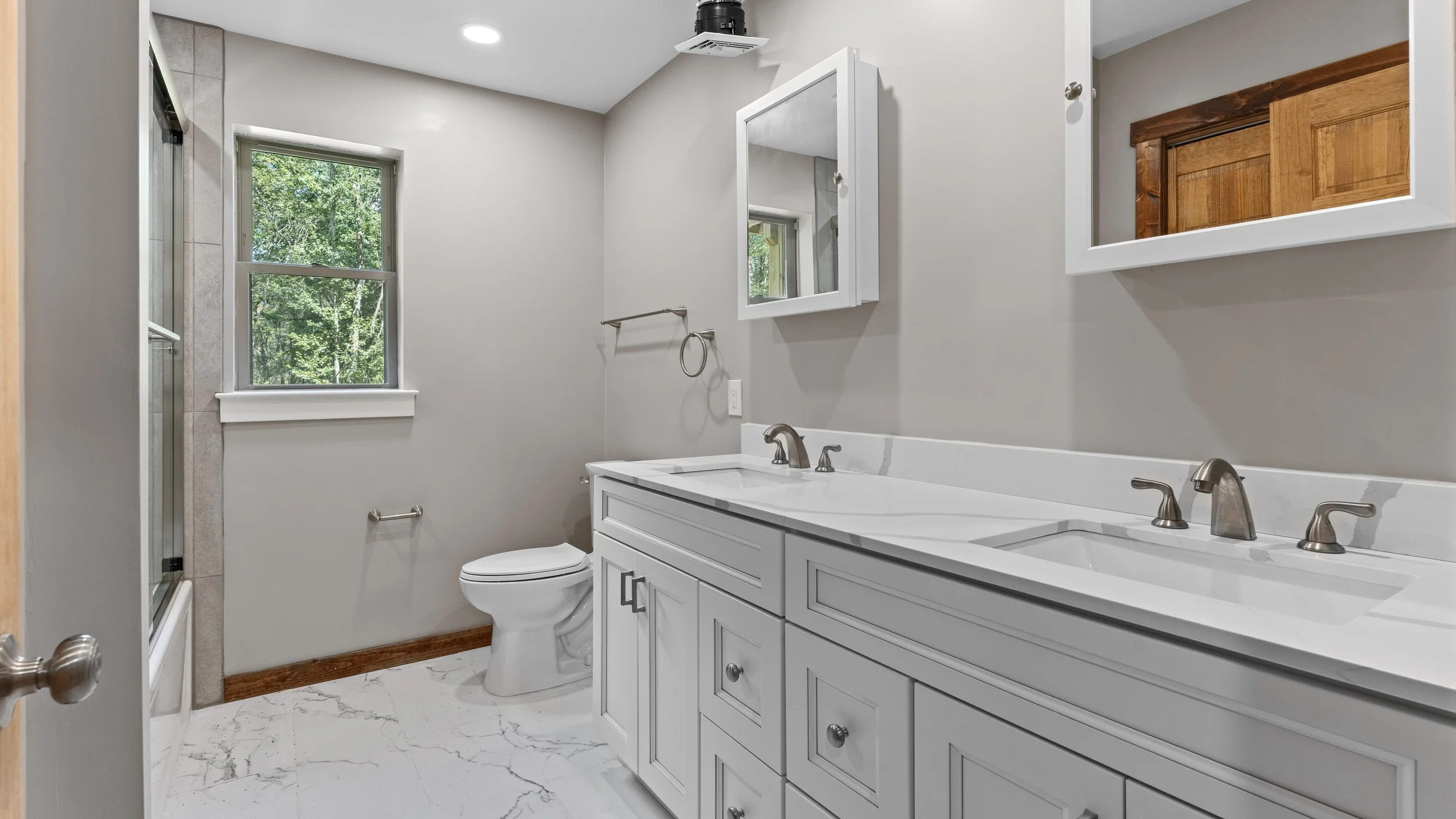 Bathroom with a double sink vanity, mirror cabinet, toilet, window with green foliage outside, marble floors, and wood accents.
