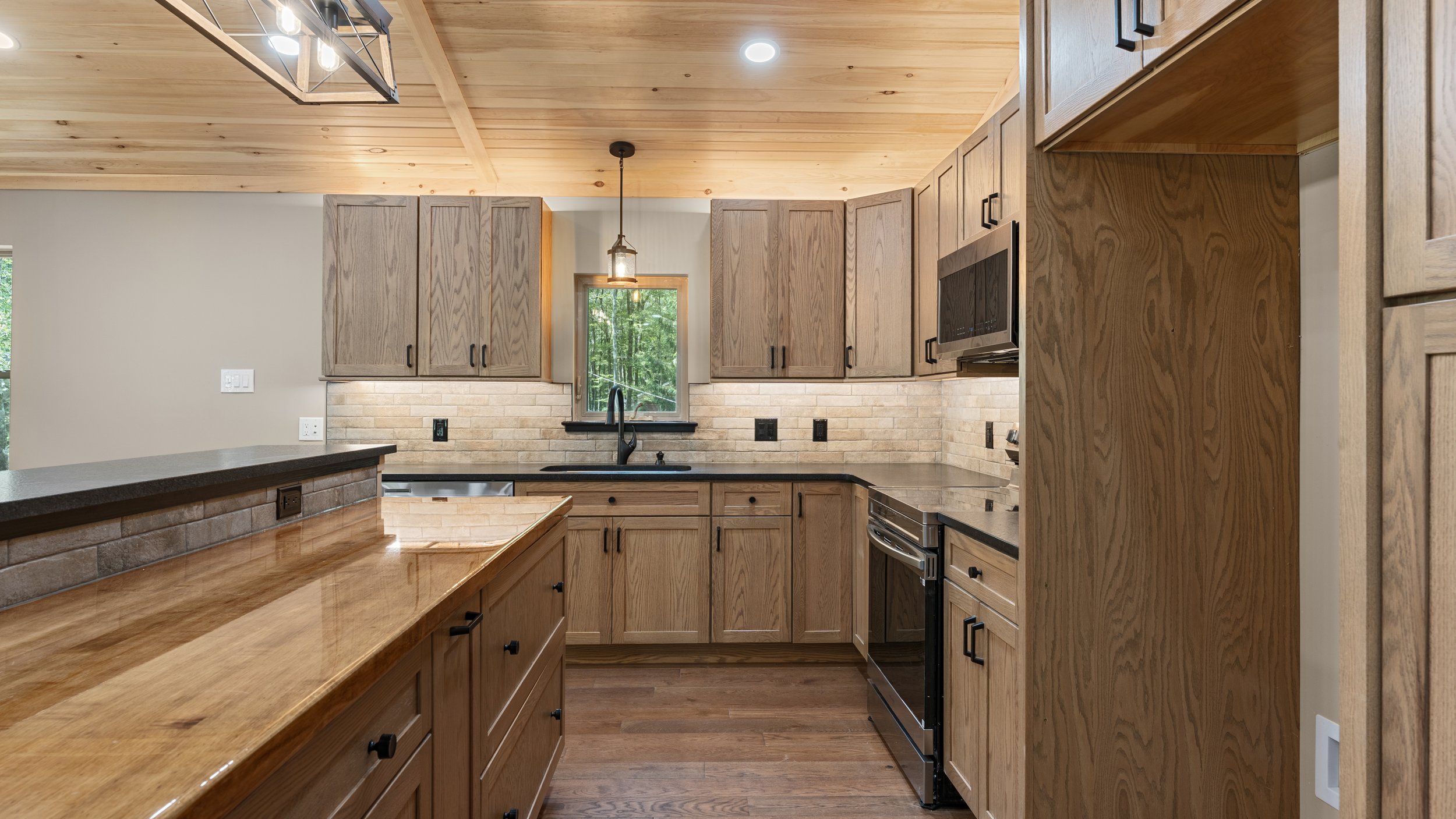 Kitchen with wooden cabinets, black countertops, brick backsplash, and stainless steel appliances, natural wood ceiling, and hardwood flooring.