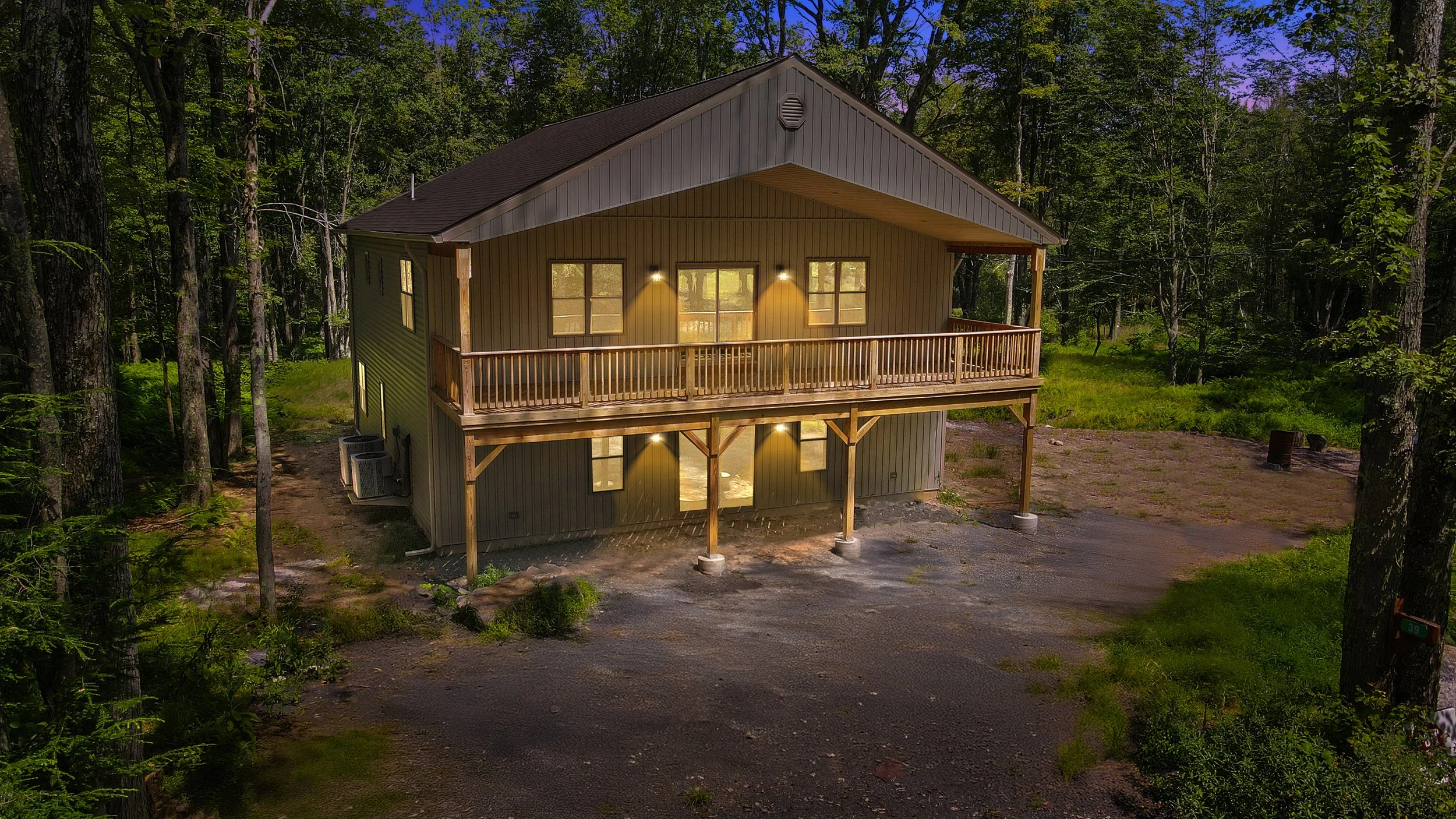 A two-story house in a wooded area with an illuminated porch and balcony at night, surrounded by trees and greenery.