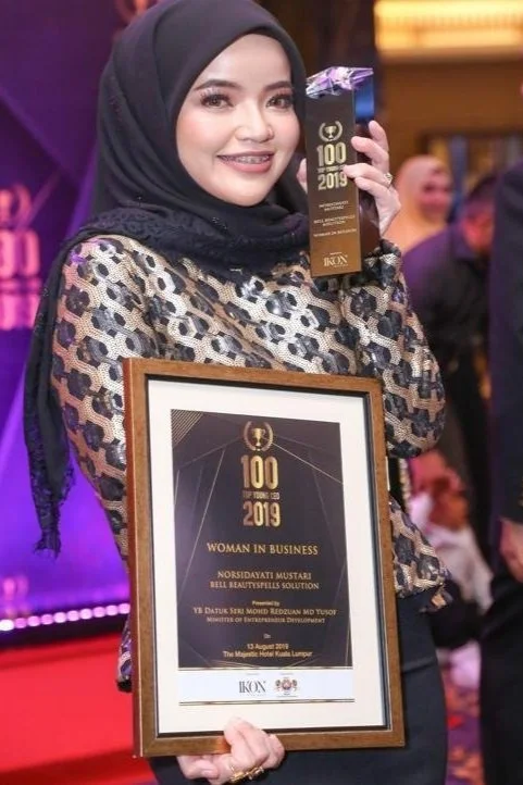 Woman wearing hijab holding a framed certificate and an award trophy at an event.