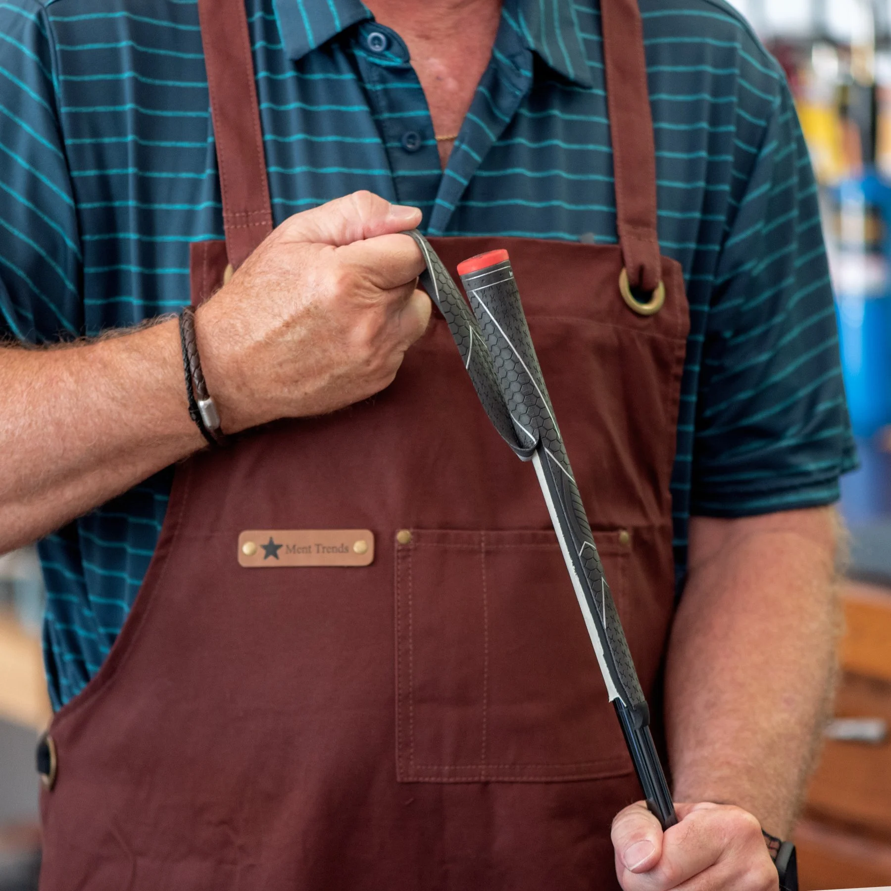 Close-up of a person holding a golf club, wearing a maroon apron over a blue striped polo shirt, regripping a golf club.