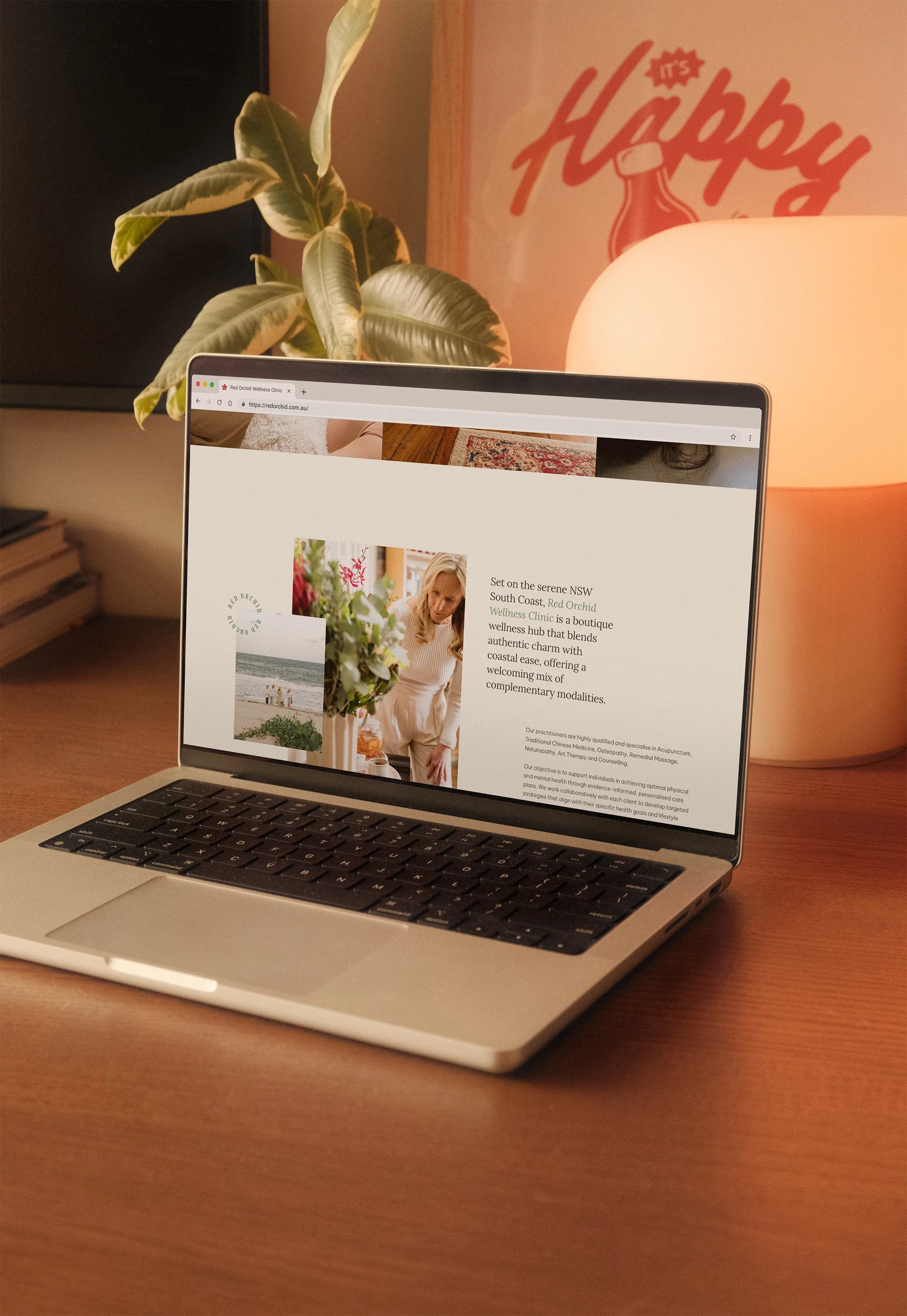 A laptop on a wooden desk displaying a webpage about Red Orchid Wellness Clinic, a boutique wellness hub located in South Coast, NSW. The desk has a large potted plant with broad green leaves, a table lamp emitting warm light, and a wall decoration with the word 'Happy' partially visible.