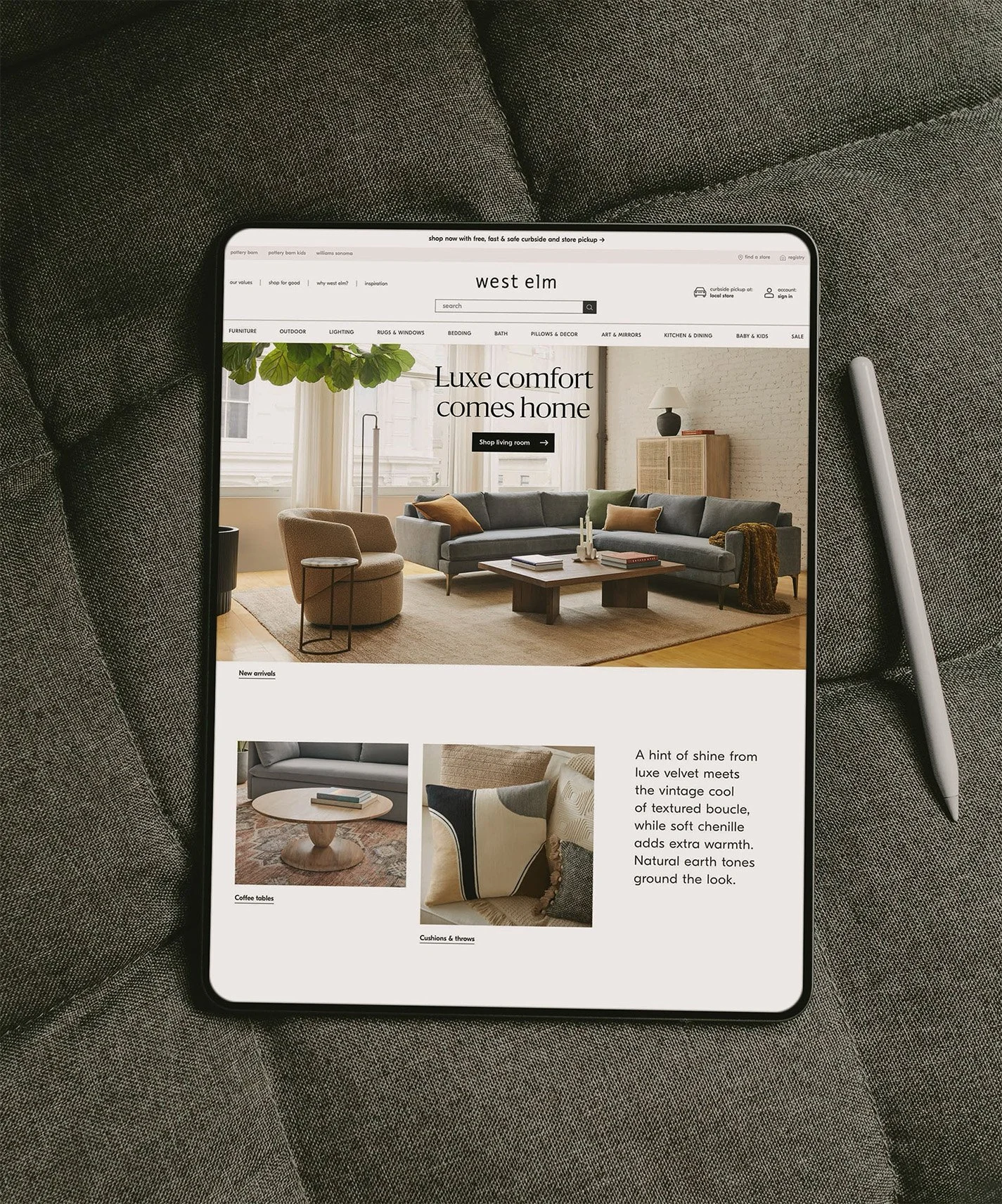 A tablet displaying a home furniture website on a gray fabric surface, with a silver stylus pen placed beside it.