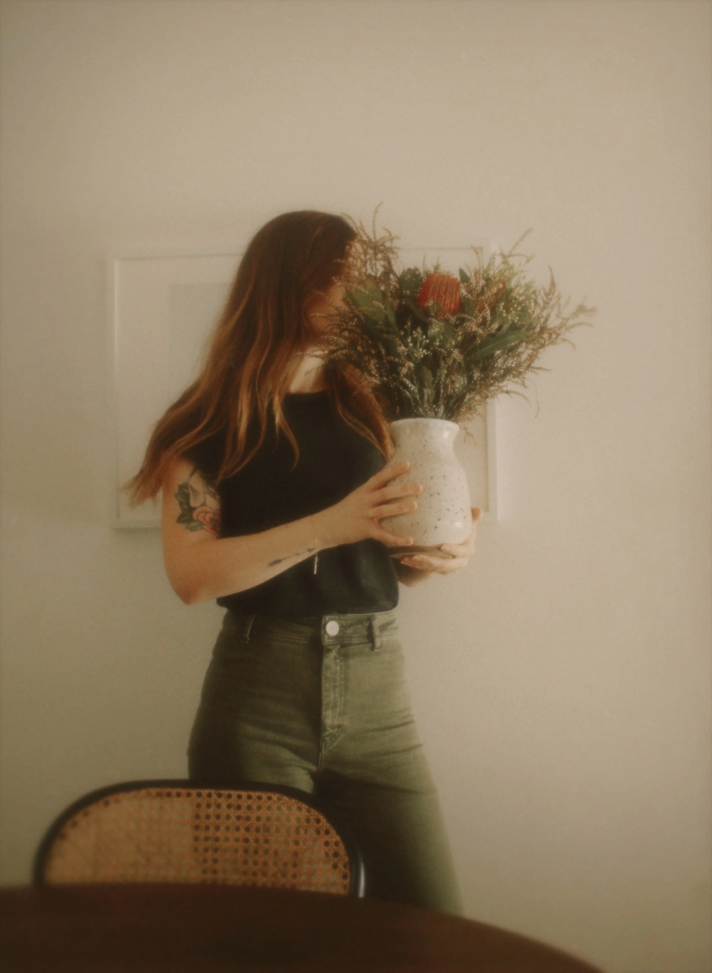 A woman with long, wavy brown hair holding a large white vase with a colorful floral arrangement, standing in front of a plain white wall.